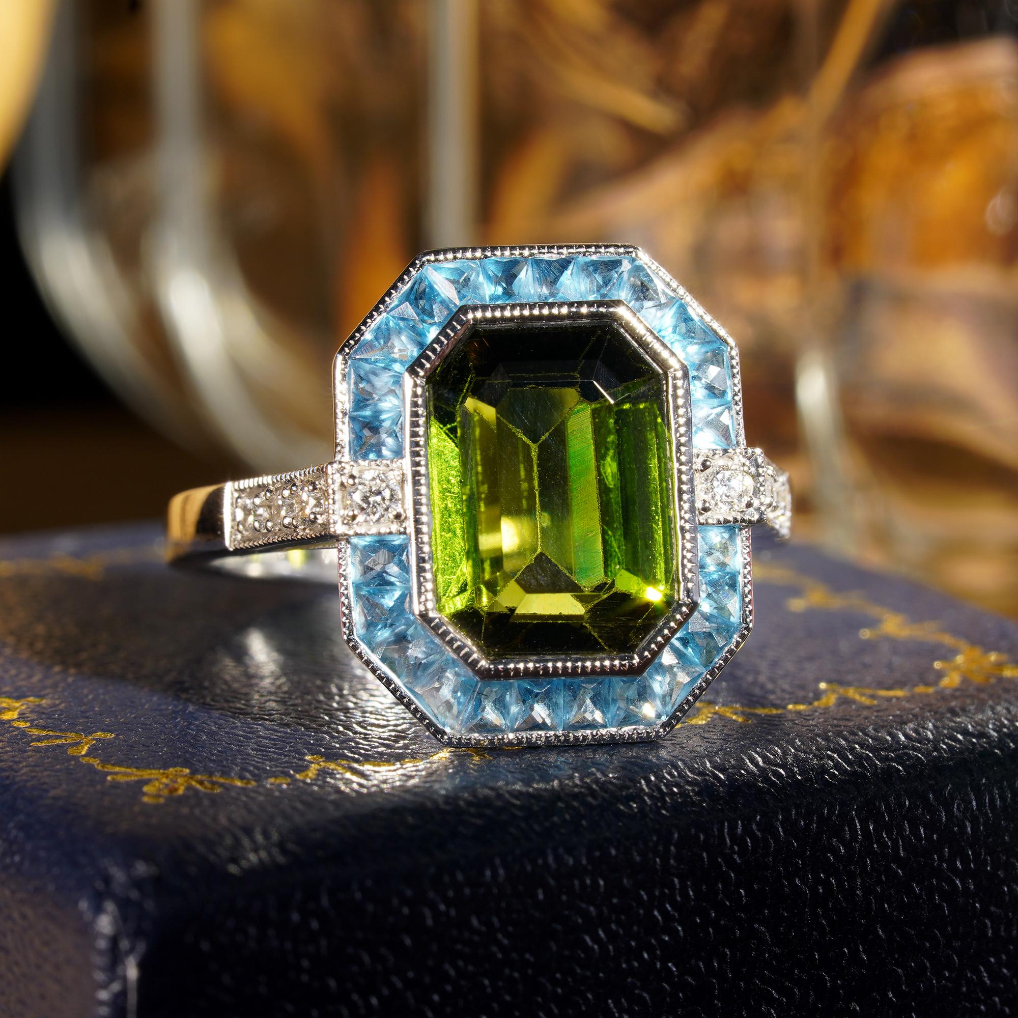 Centering an emerald cut green tourmaline, accented by French cut London blue topaz, enhanced by round cut diamonds. This Art Deco inspired ring is a perfect piece for an engagement ring, or to commemorate an anniversary.

Ring Information
Style: