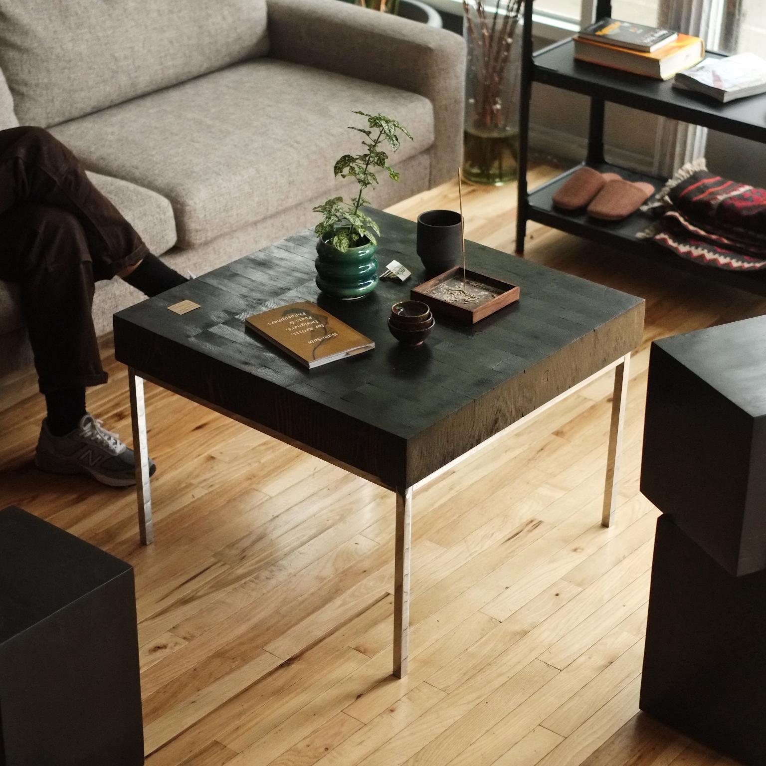 The Fleur Noir coffee table is a haptic sculpture. The coffee table's top is a 'butcher block' style heavy slab of blocks of pine.
Each end grain piece exposes a kerf cut to the table's surface top to the senses of touch and sight. The select