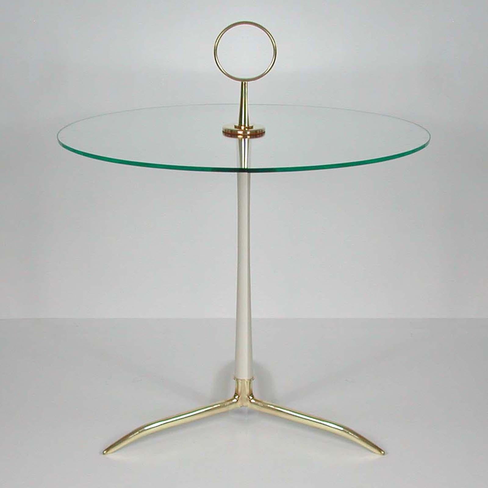 This elegant midcentury side table was designed by Cesare Lacca and manufactured in Italy in the 1950s. 

It is made of brass, light cream lacquered metal and clear glass with a polished edge on a tripod base. The round handle makes it easy to