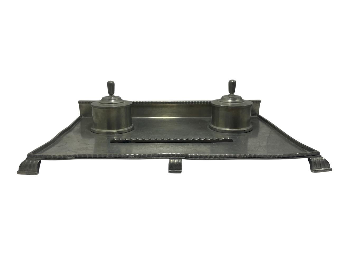 Elegante calamaio doppio realizzato dal famoso argentiere svedese CG Hallberg, risalente al 1931. Realizzato in peltro riccamente patinato e martellato a mano, il pezzo presenta una base rettangolare a vassoio con due calamai cilindrici e