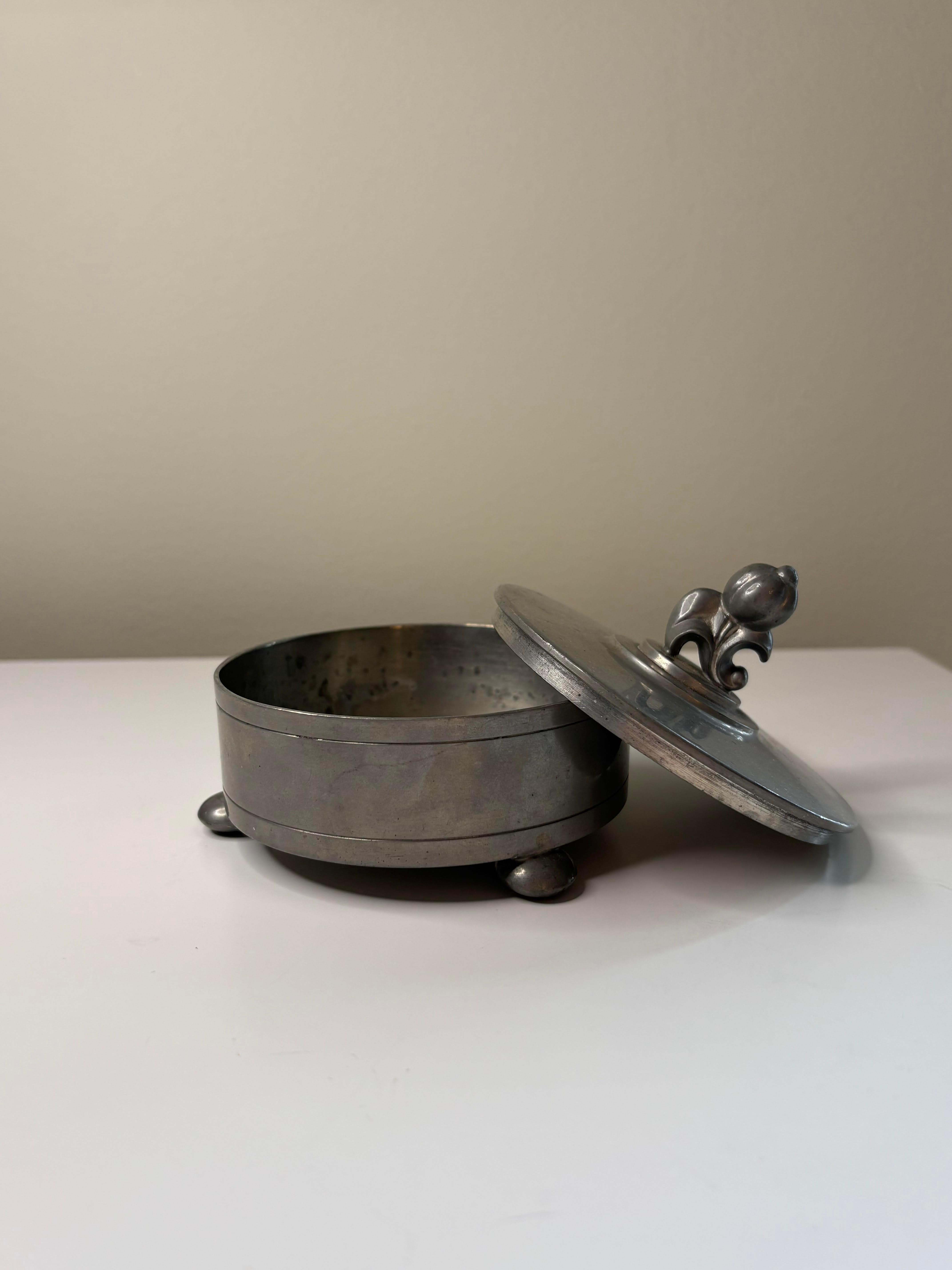 Elegante scatola con coperchio in peltro realizzata dal famoso argentiere svedese CG Hallberg nel 1930. Caratterizzato da una forma arrotondata con piedi a sfera e un terminale scultoreo, questo pezzo mette in mostra la raffinata maestria