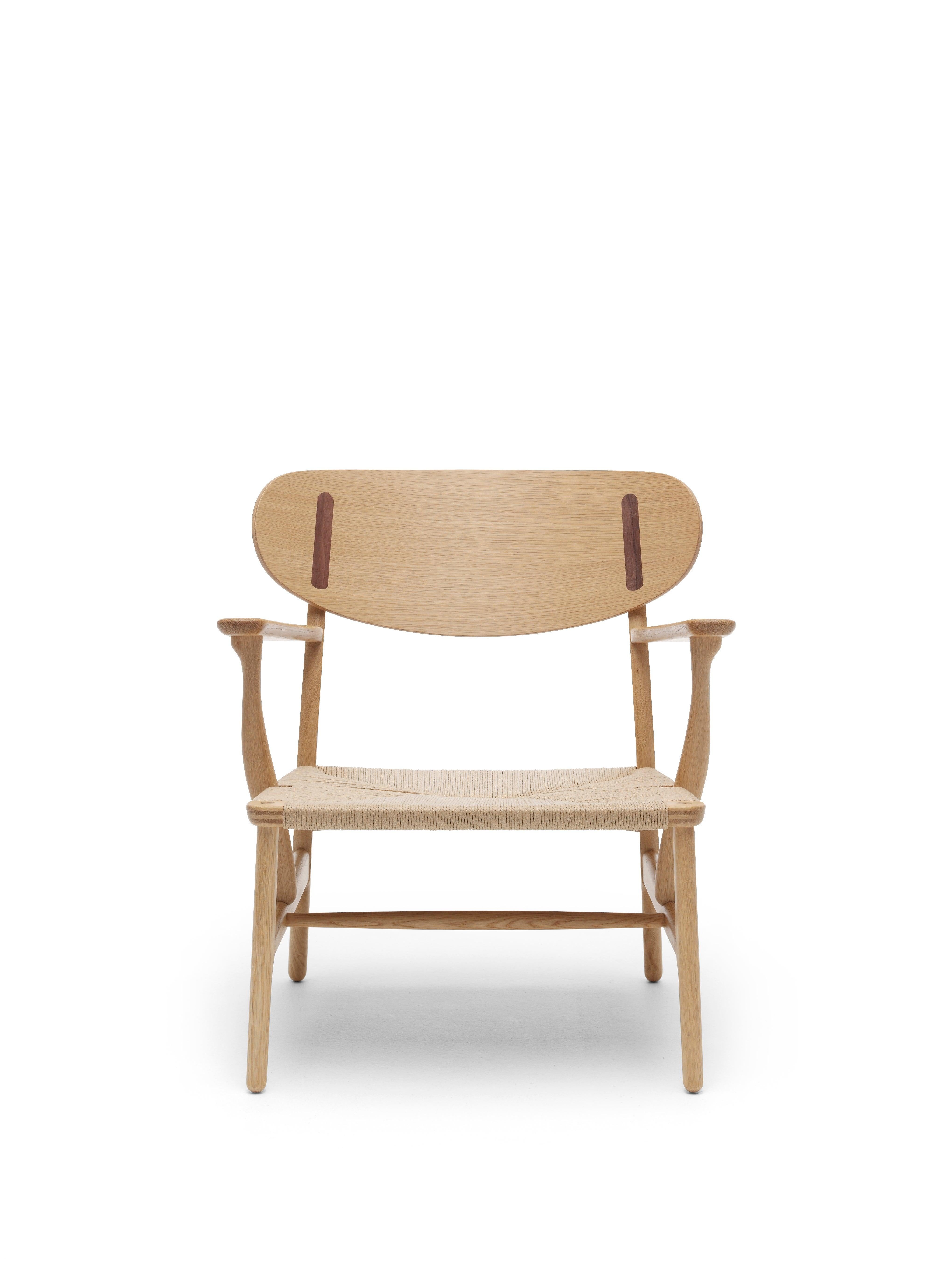 The CH22 lounge chair by Hans J. Wegner was brought back into production by Carl Hansen & SÃ¸n in 2016. It features elegant armrests, an envelope-woven paper cord seat, and a distinctive back shell in form-pressed veneer with striking oblong cover