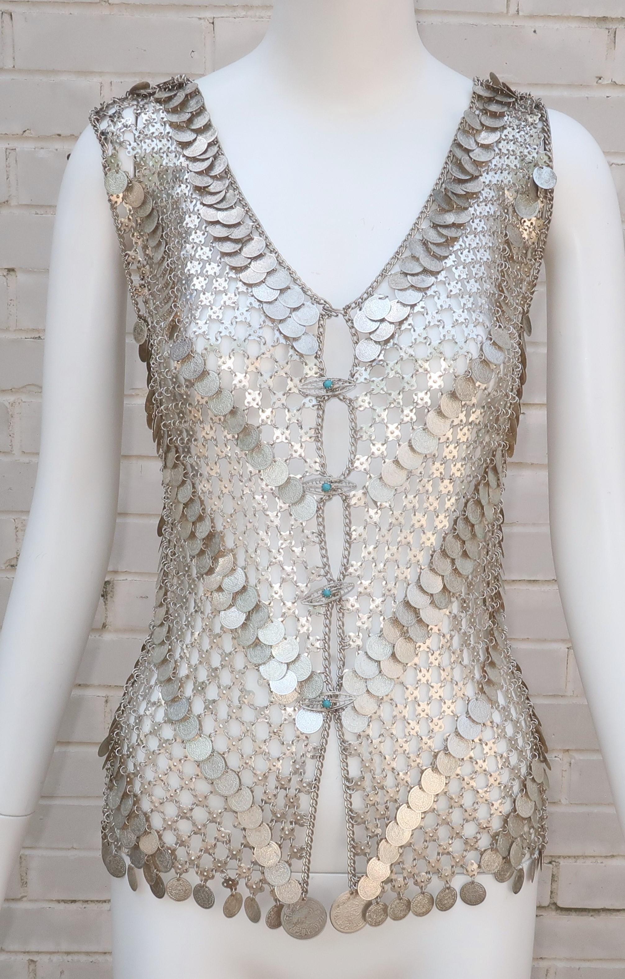 Put on your fashion armor with this 1960's silver metal chain link vest with coin embellishments throughout.  The v-shaped vest closes at the front with a top hook and diamond shaped metal 'buttons' accented by turquoise glass beads.  The exotic
