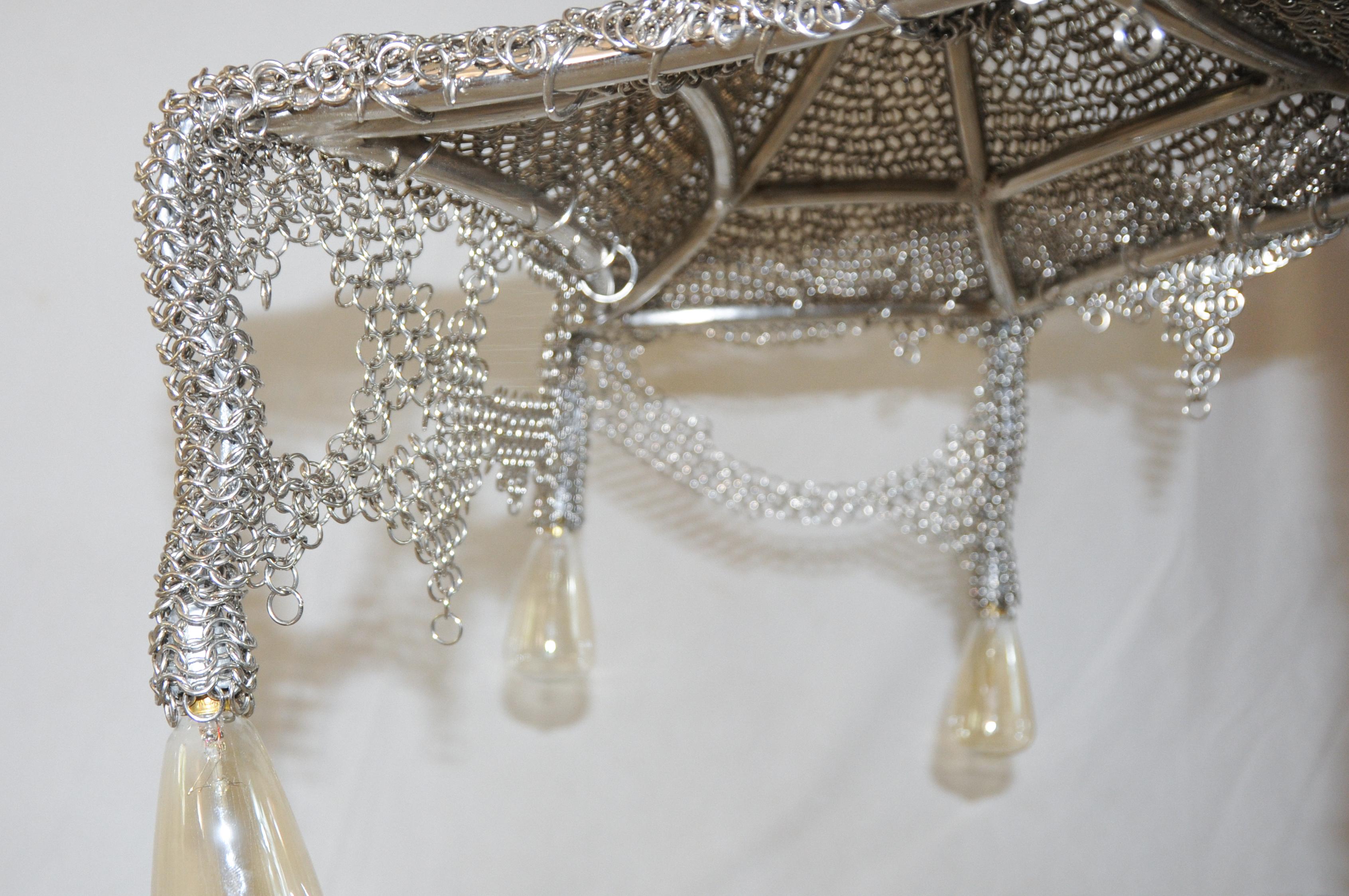 Fusione di artigianato medievale e moderna eleganza gotica, il lampadario Chainmail Cobweb evoca la bellezza eterea di una ragnatela. Meticolosamente realizzato a mano con anelli di metallo intrecciati, il suo velo di cotta di maglia a cascata