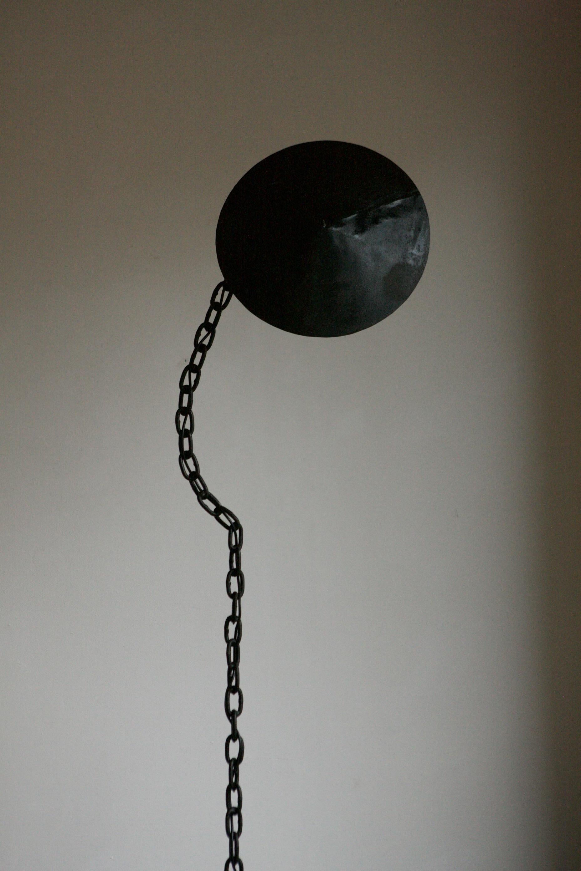 A black chain floor lamp with a cone shade that shields a bulb holder. The chain stem is welded together in a fluid form to give the illusion of a hanging chain.
