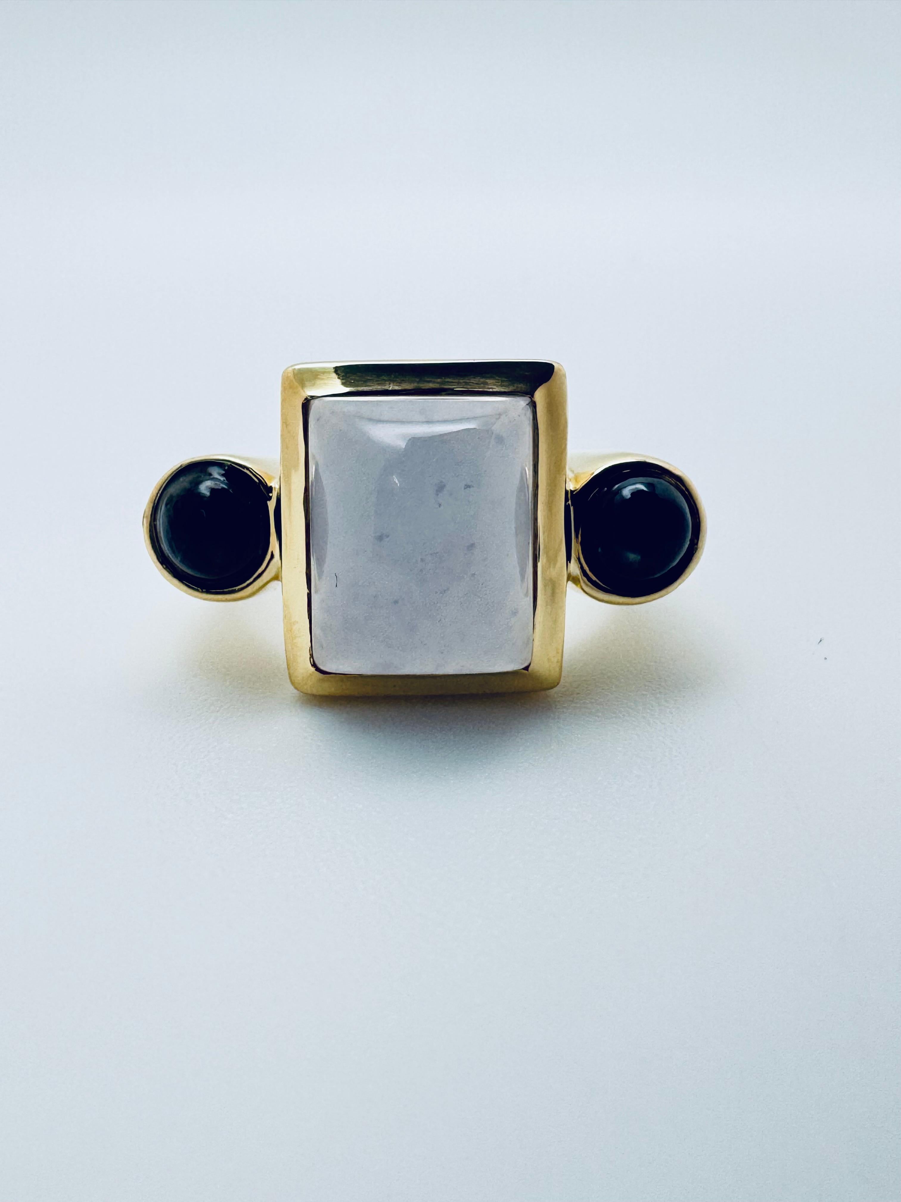 Cette magnifique bague présente un mélange captivant d'élégance douce et de design audacieux. Réalisé en luxueux or jaune 18 carats, il est orné d'une calcédoine lisse et polie qui rayonne d'un éclat serein et translucide. Deux saphirs d'un bleu