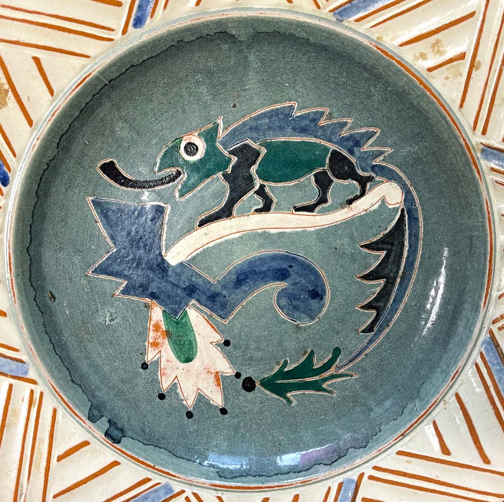 Beautifully designed and glazed, this striking, large bowl depicts a blue and green chameleon at its center that is climbing a highly stylized Art Deco foliate element, framed by a wide, flat rim decorated with a complex, geometric chevron pattern.