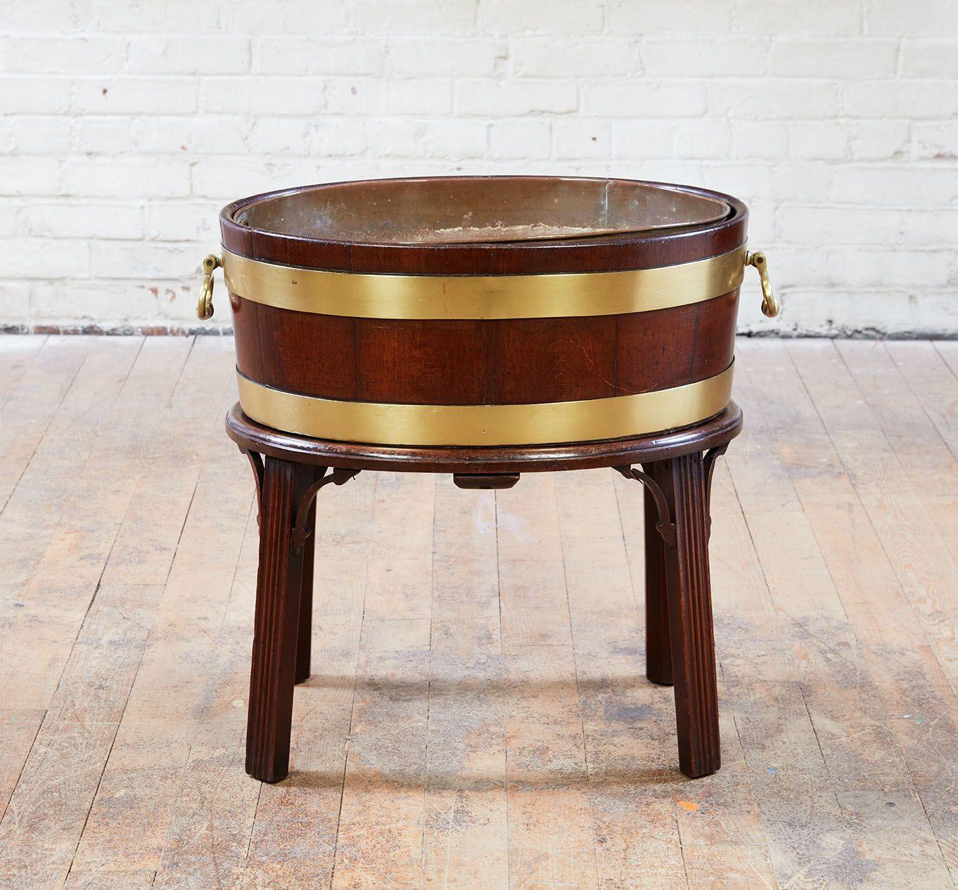An 18th century English wine cooler on stand, featuring coopered mahogany basin with brass banding and original copper liner.  Well-formed solid brass carrying handles.  All on mahogany stand with slightly splayed legs and fretwork corner brackets. 