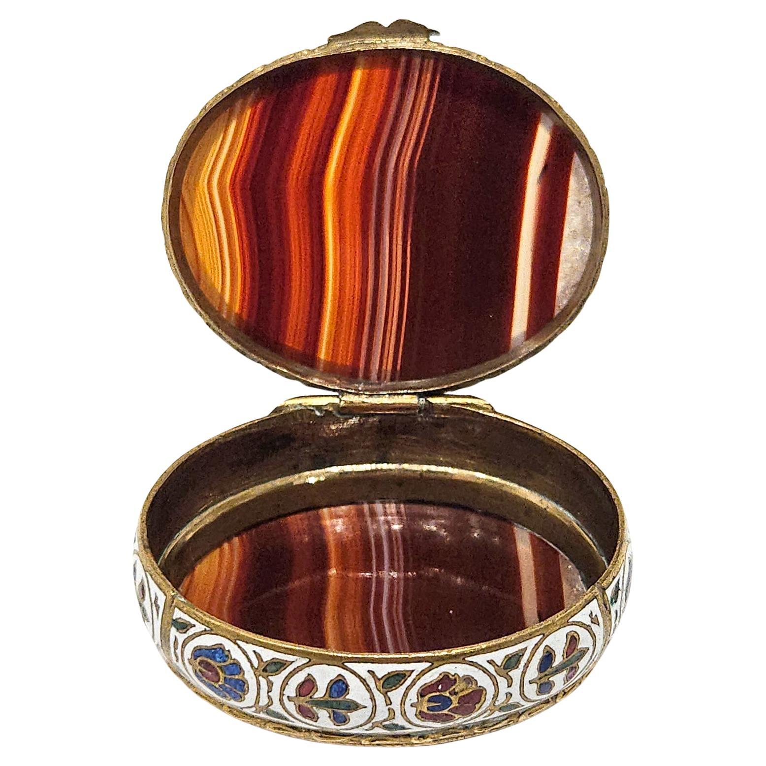 An elegant and very collectible 19th century French gilt metal, champlevé enamel and banded agate pill or snuff box made c. 1880.

The box has oval form with a frieze of colourful flowers described in champlevé enamel against a white ground that