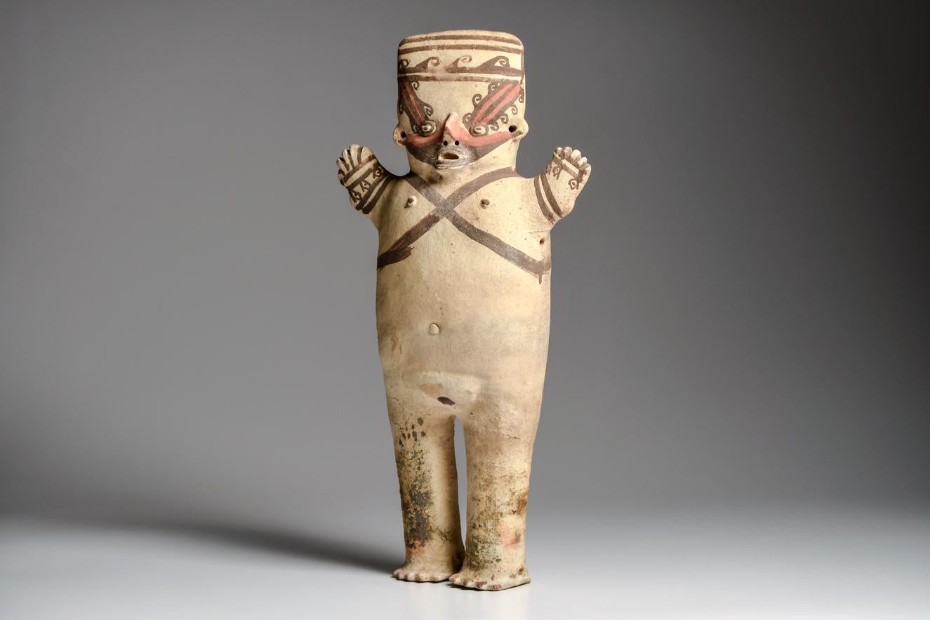 Large standing female figure with raised arms. Relief facial features, with red-orange and chocolate brown painted decoration, on a cream slip ground. Crossed straps on her chest, and geometric decoration on her arms and back of both legs. Repaired