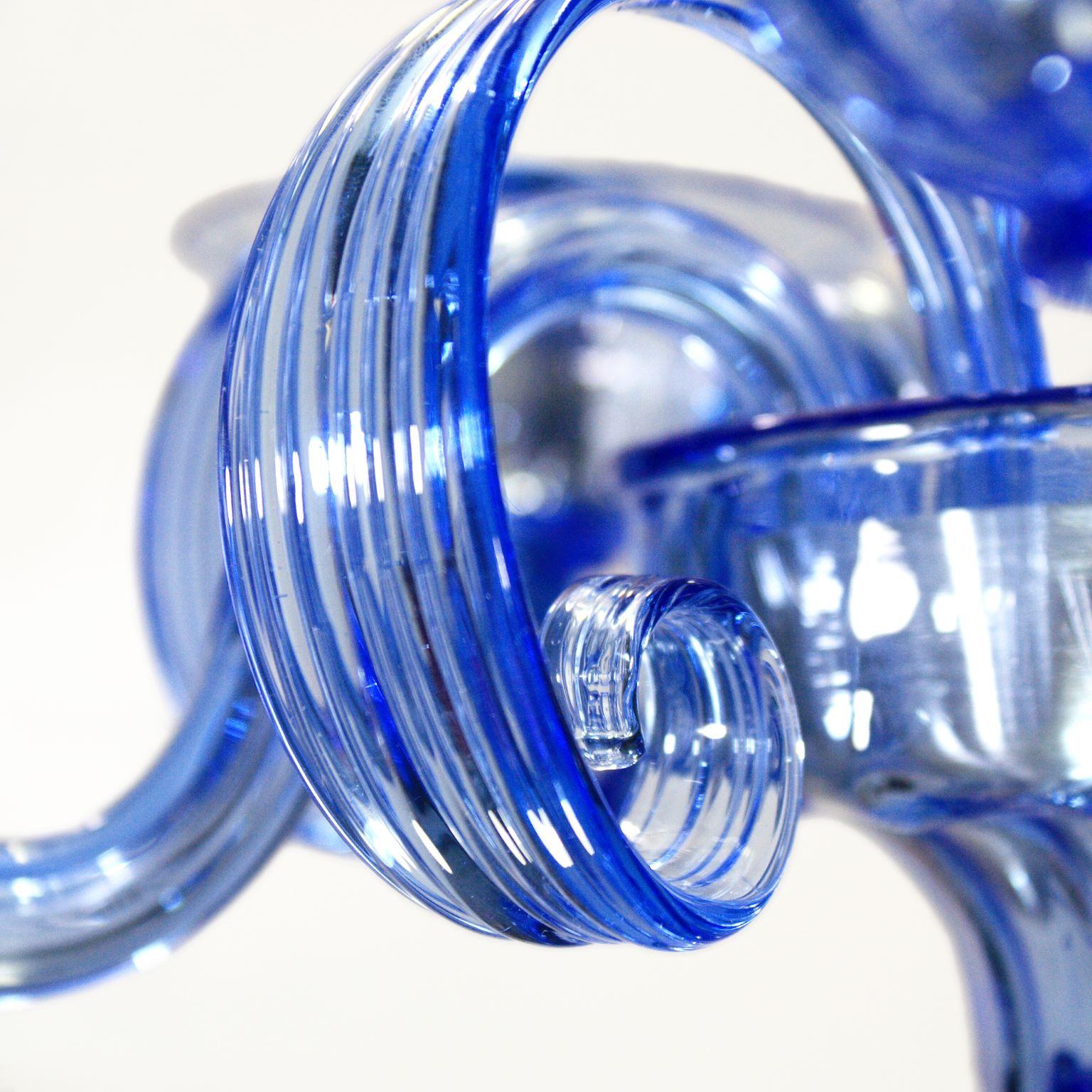Capriccio Kronleuchter, 5 Lichter, blaues Kunstglas, mit geschwungenen Ornamenten von Multiforme.
Es ist von der klassischen venezianischen Tradition inspiriert und zeichnet sich durch eine zentrale Säule aus, in der zahlreiche 
