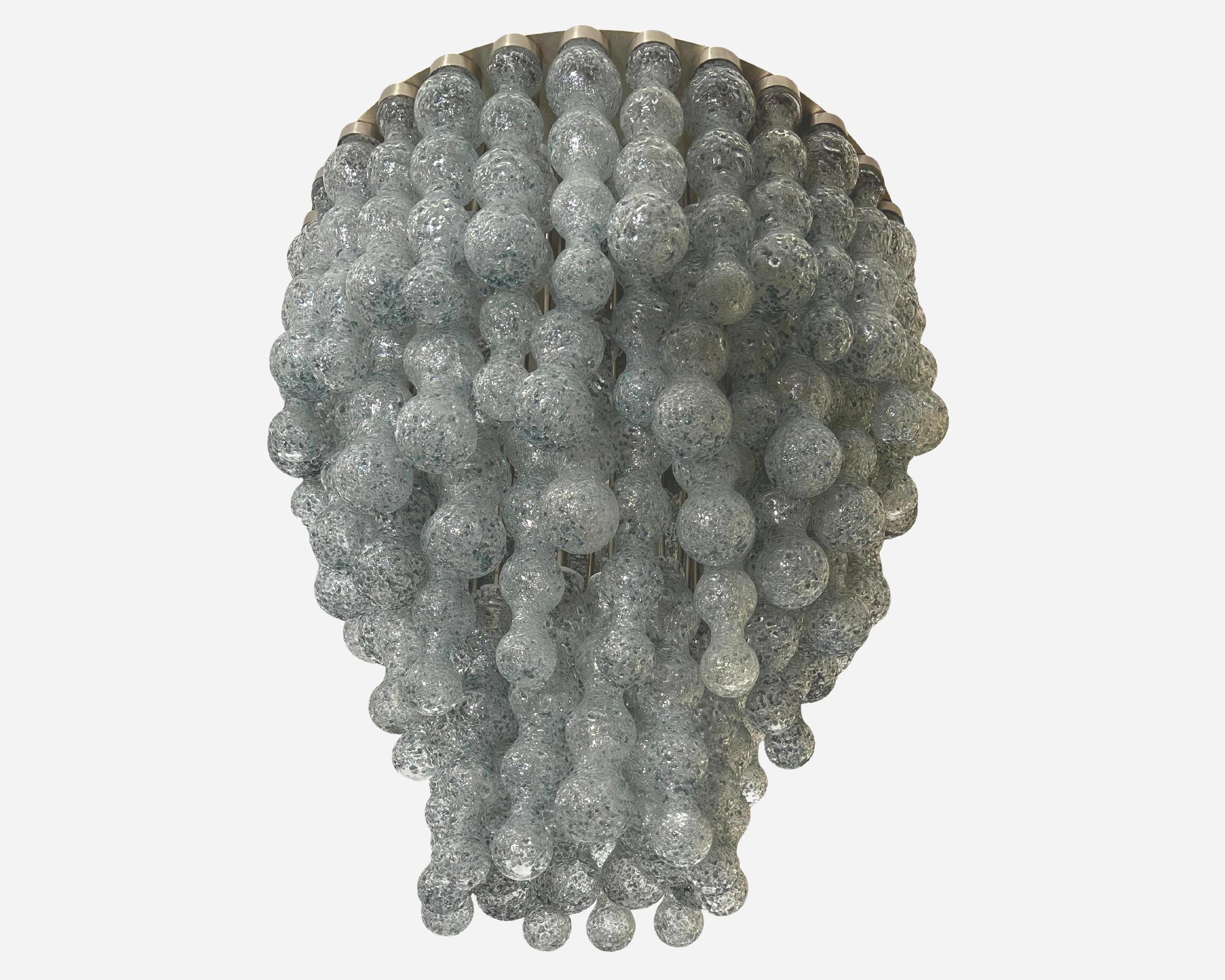 Interesante lámpara de techo hecha con piezas de vidrio soplado a las que se han pegado con calor un gran número de escamas de vidrio gris azulado.
Las piezas de cristal ocultan cuatro bombillas. La placa base es de acero inoxidable, al igual que