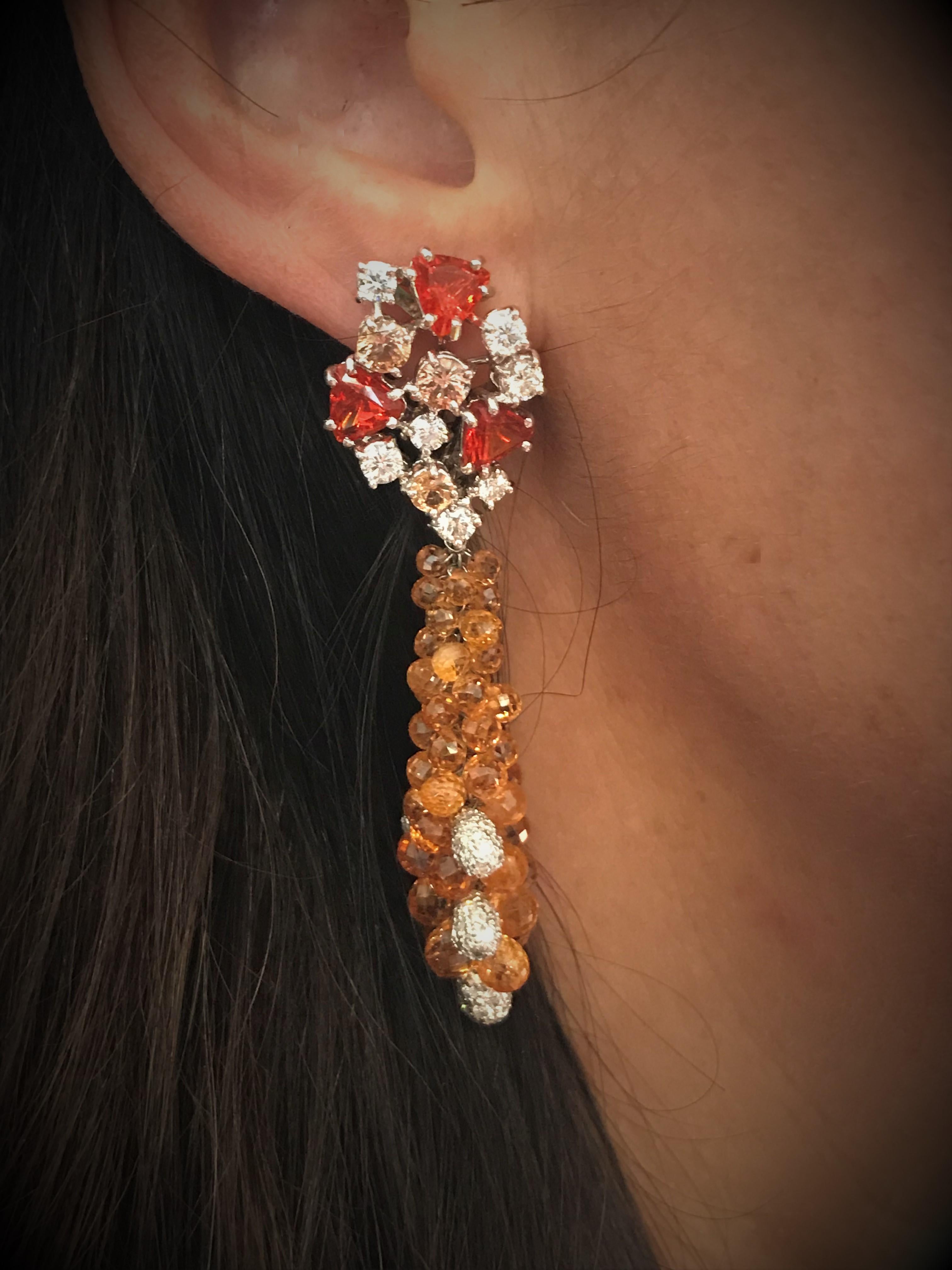 The warm color of the orange briolette cut sapphire are underlined by sparkling diamonds in these charming earrings.
White gold, brilliant pears, briolette cut spessartines, irregular red opal fires, round brilliant cut diamonds and yellow
