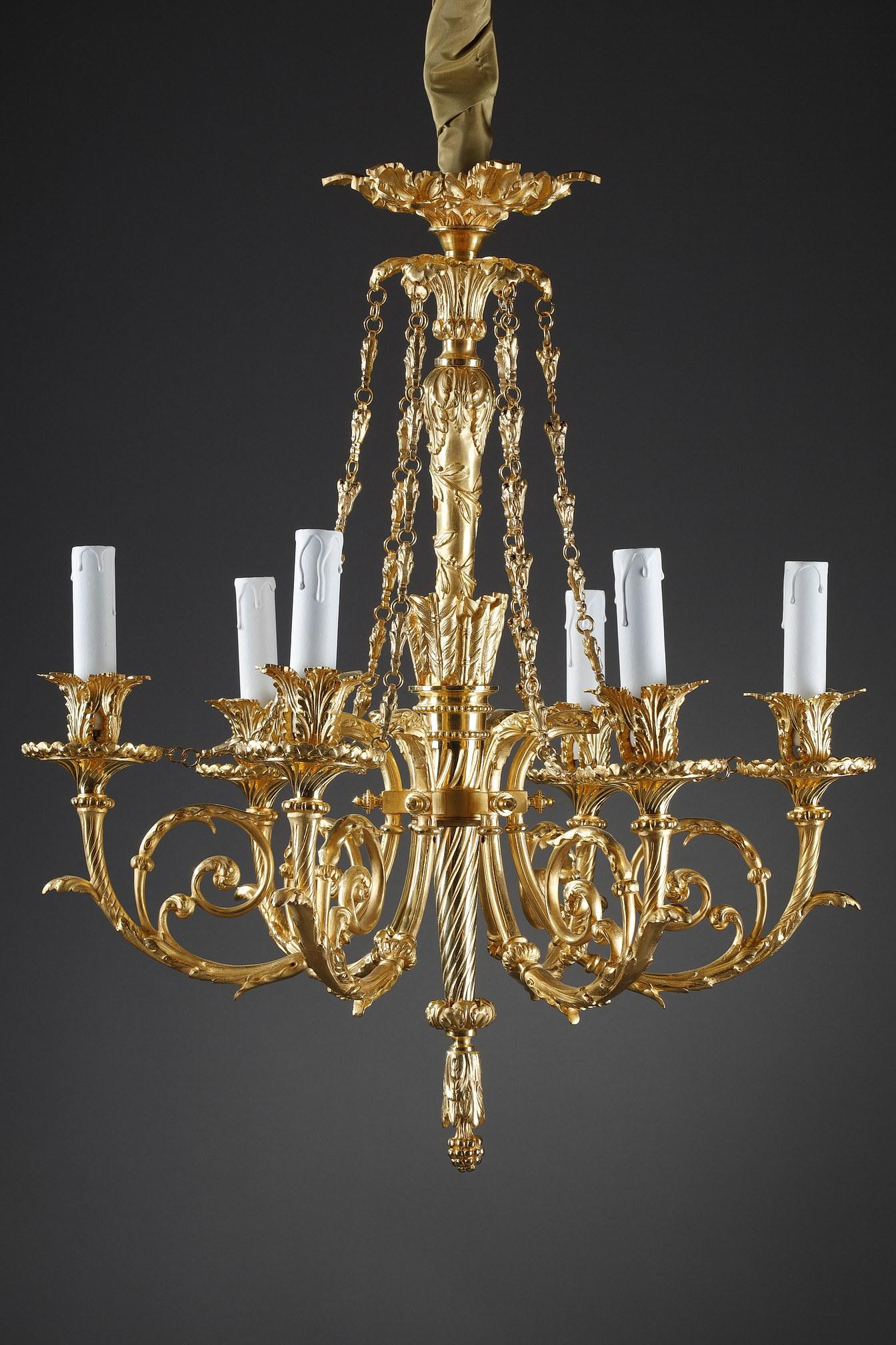 A Louis XVI style chandelier in gilt and chased bronze with 6 arms of light. The shaft in the shape of a quiver filled with arrows is prolonged by a scroll of olive branches and ends in the lower part by a pine cone. The medallion is in the form of