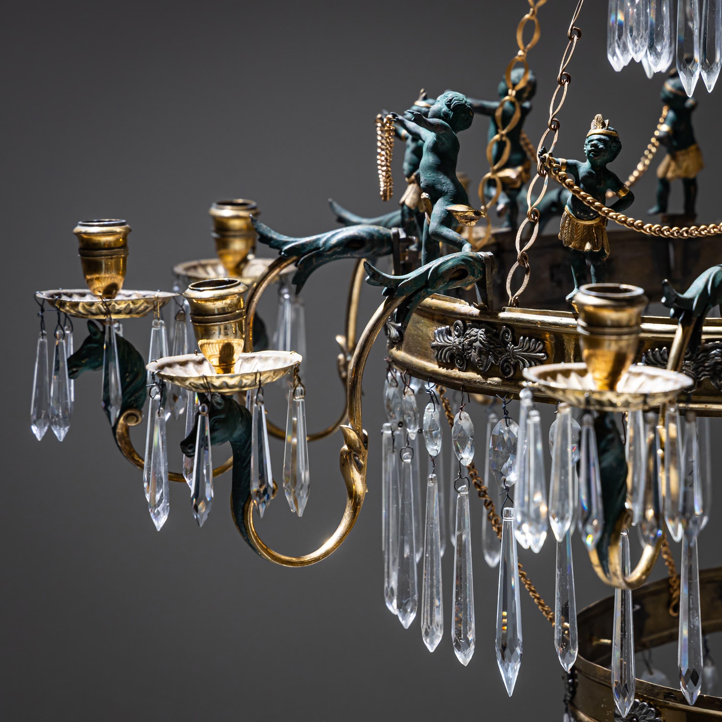 Magnifico lampadario a dodici luci con quattro livelli, sormontato da una corona di girasoli stilizzati. Realizzato in bronzo e prismi di vetro tagliato, l'anello più largo è ornato da un fregio continuo di piccoli putti, alcuni patinati verdi,