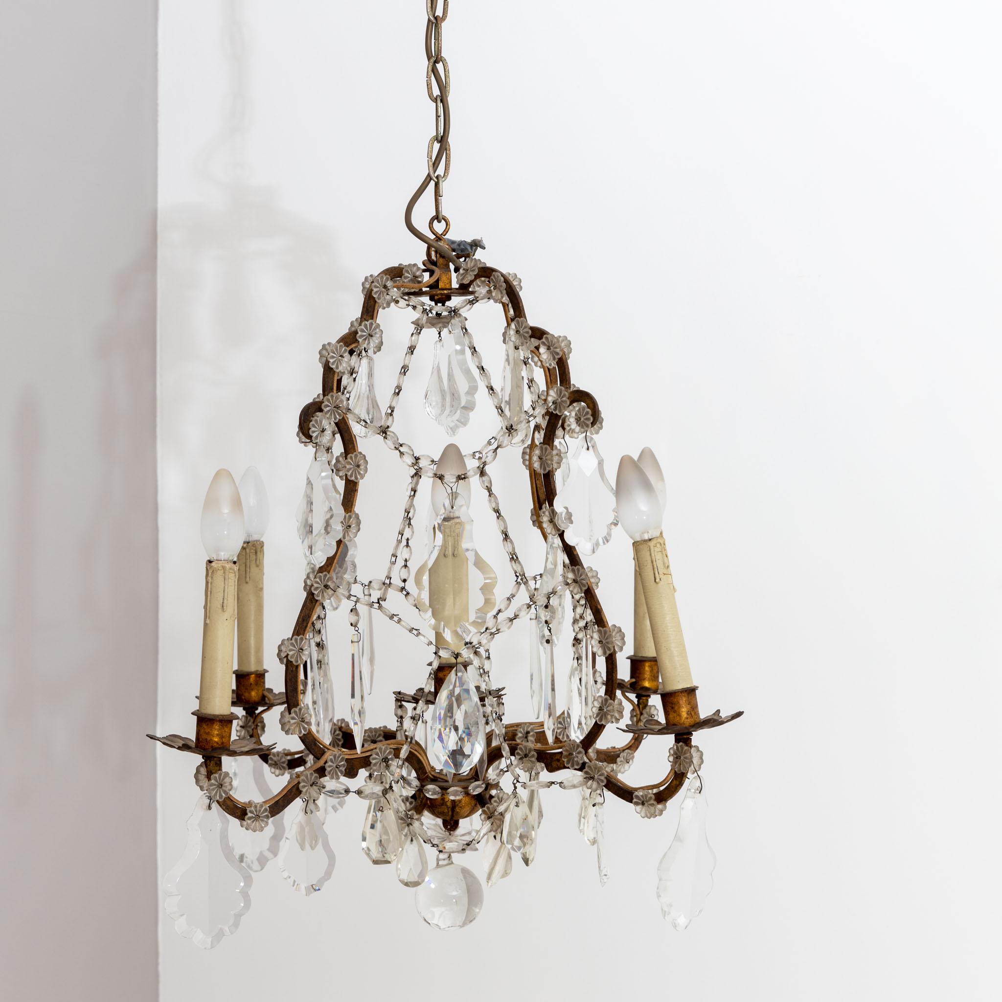 Chandelier made of patinated brass with glass prisms and five sockets in the shape of stylised wax candles. The glass hanging consists of cut prisms, partly oval and partly in leaf shape, small flowers as well as a spherical pendant in the centre.