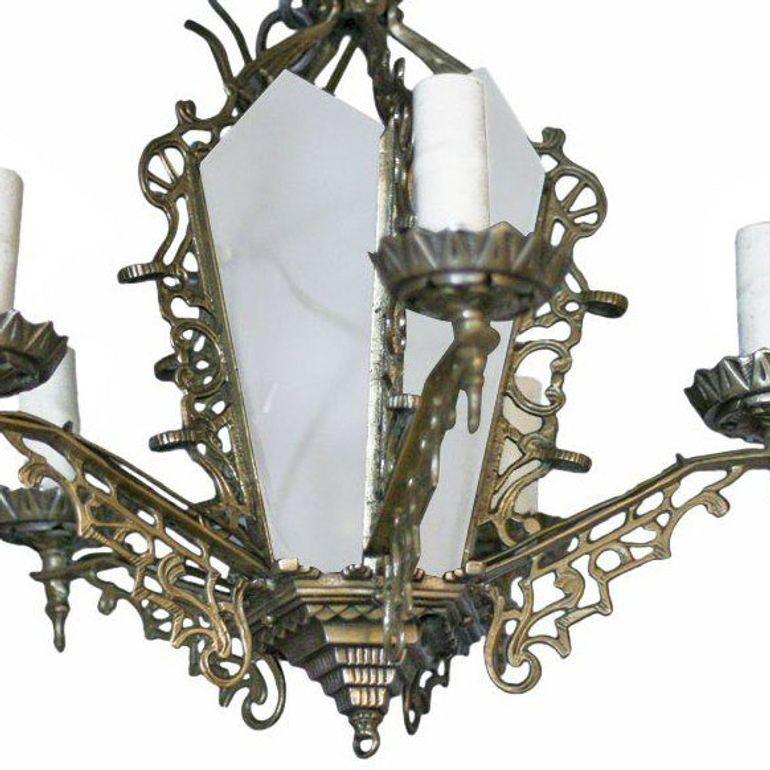 The six arms of this chandelier extend from the base of a glass paneled pendant. The piece features scrolling details throughout while the base of the chandelier features a geometric pattern.