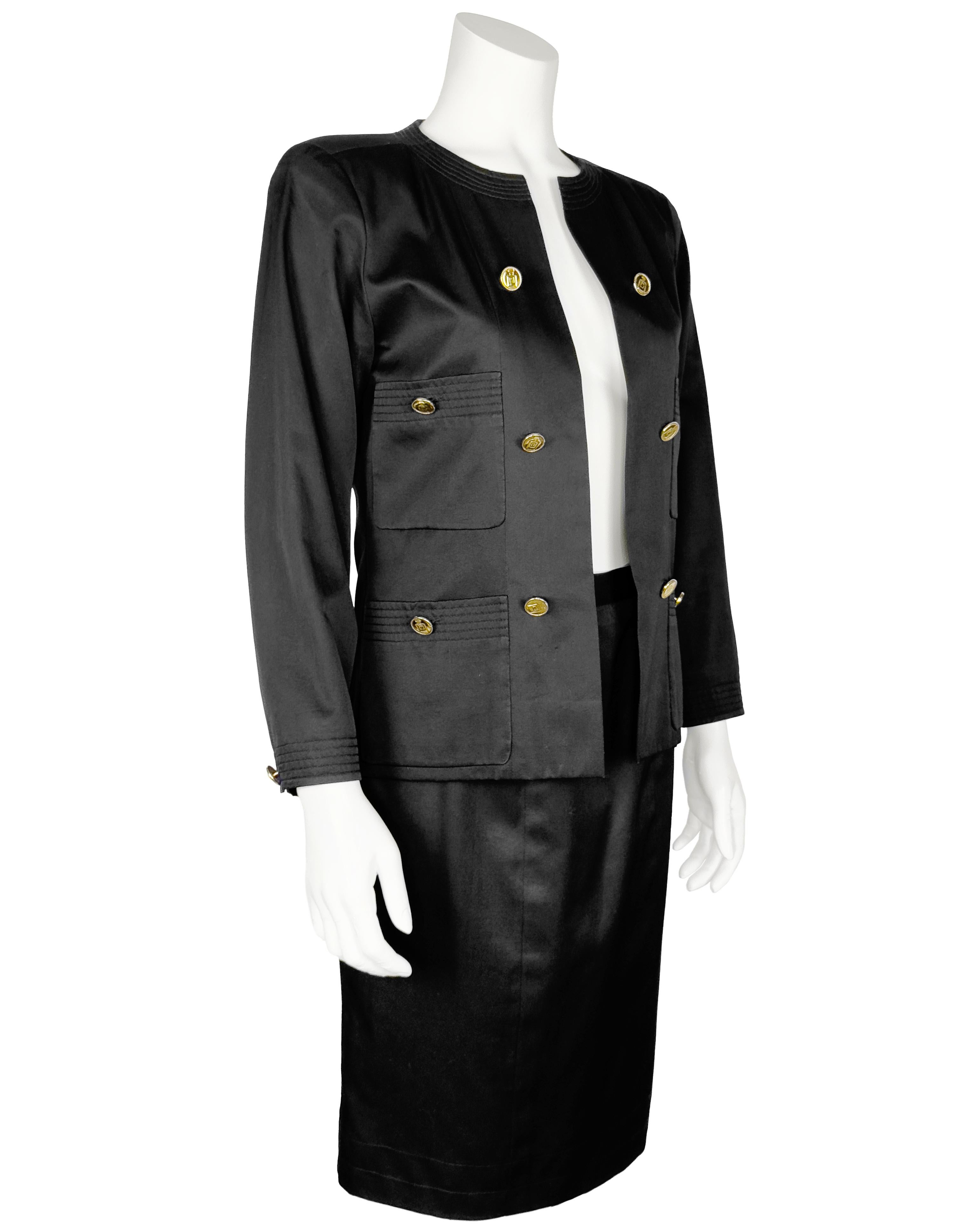 Tailleur jupe en coton noir poli des années 1980 de Chanel, avec boutons de bouteille de parfum en métal doré le long des panneaux avant, sur les quatre poches plaquées et sur les manches. Veste ouverte sur le devant, doublée de soie noire portant