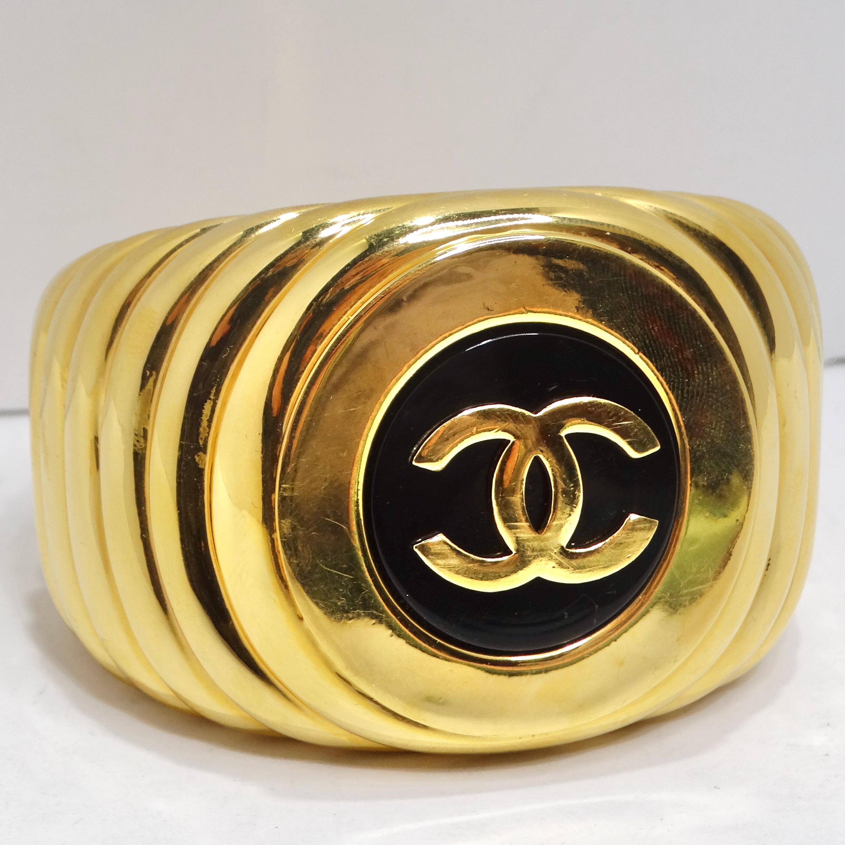 Elevate your style with the Chanel 1980s CC Logo Ribbed Gold Tone Cuff Bracelet – a beautiful statement cuff that captures the essence of vintage Chanel glamour. This cuff features yellow gold plating with a ribbed effect, gracefully giving way to a