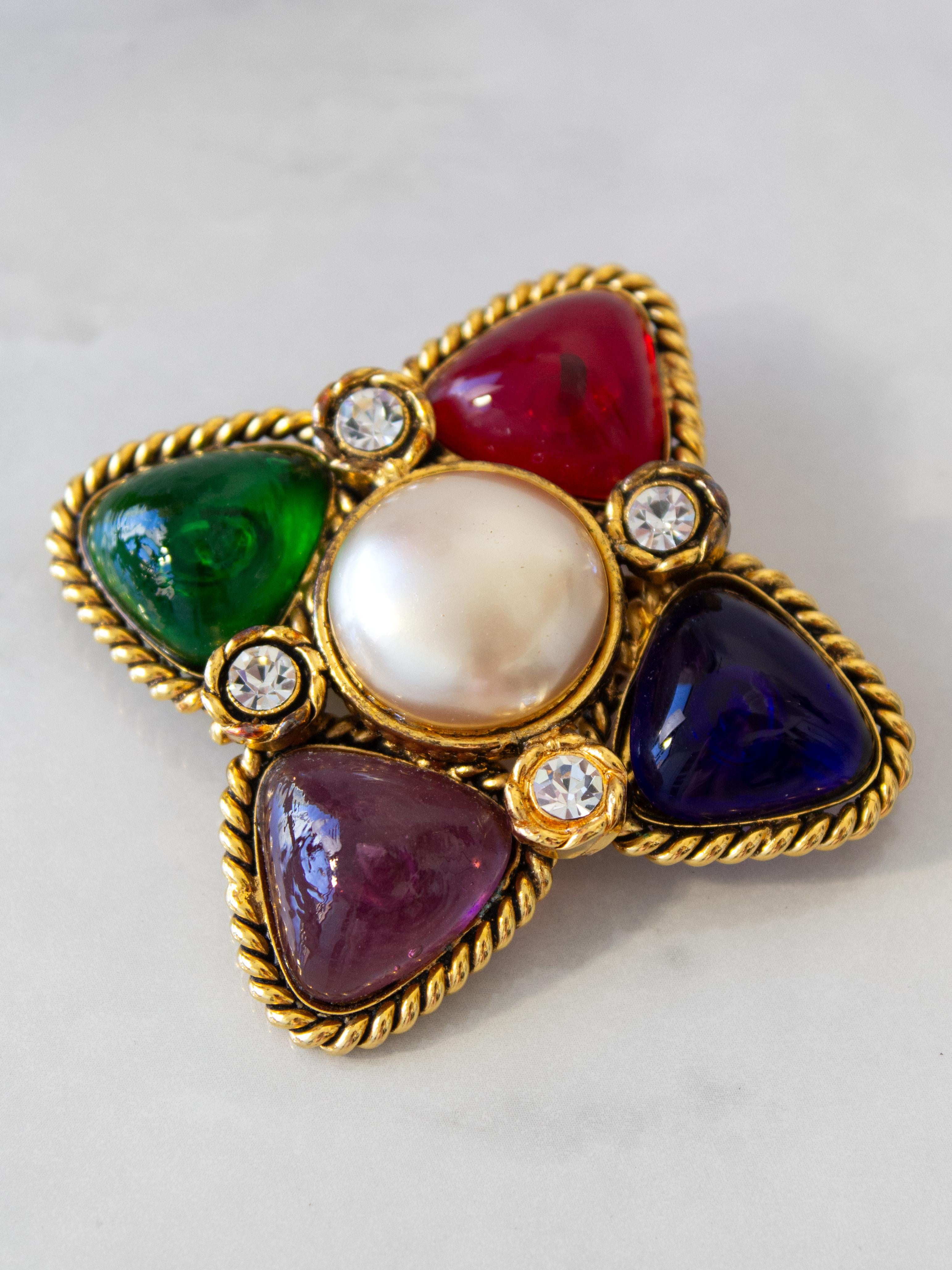 This exquisite vintage Chanel brooch from collection 23 exudes timeless elegance with its gold-tone braided gilded metal and multicolored red/blue/purple/green glass paste stones. The captivating cross rhombus motif, adorned with center faux pearl,