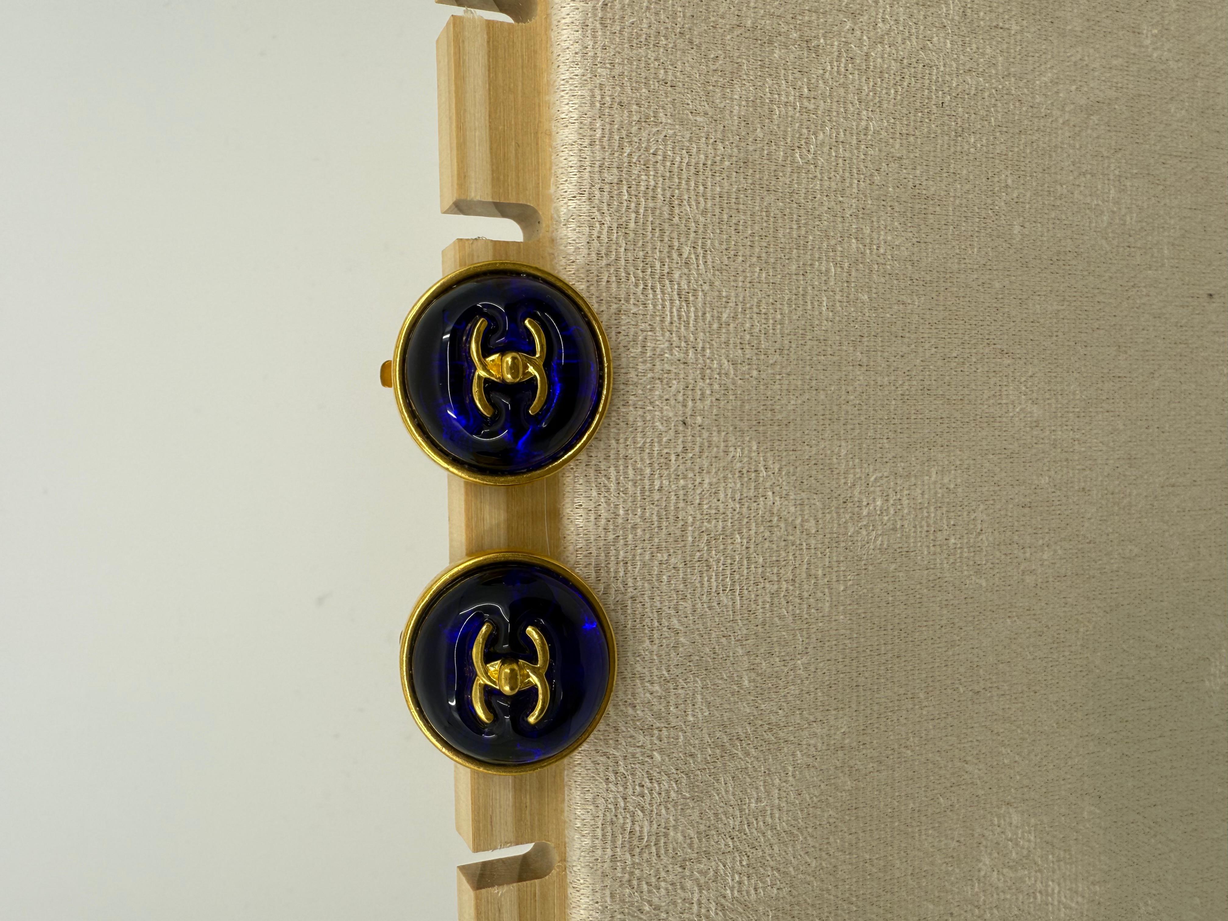 Un exquisito par de pendientes de clip Chanel vintage de 1997, con centros de cristal vertido (Gripoix) de color azul cobalto encerrados en monturas redondas doradas. En el corazón de cada pendiente se encuentra el icónico emblema del cierre