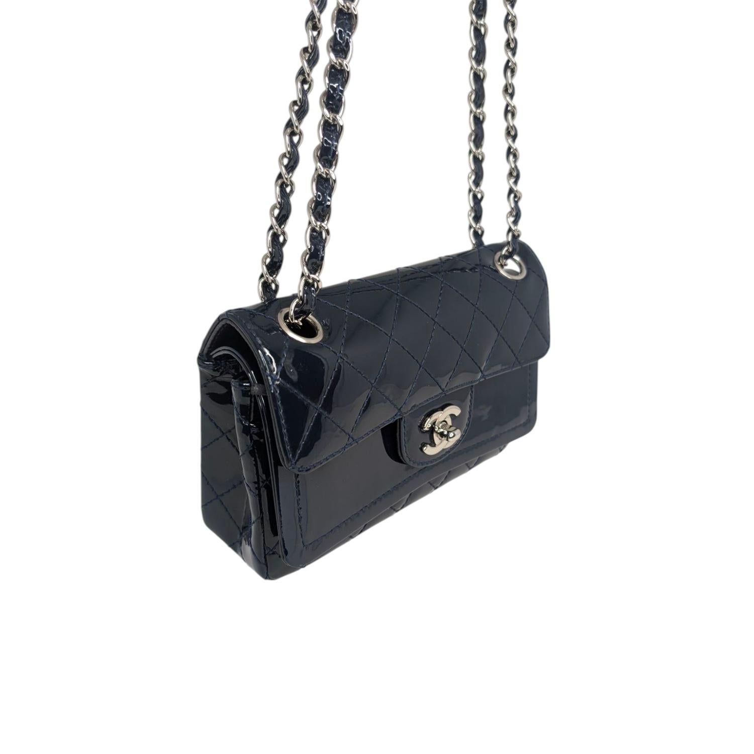 Este elegante bolso bandolera está confeccionado en charol acolchado con brillantes rombos en color azul. El bolso tiene un bolsillo trasero de parche, una correa bandolera de eslabones de cadena de color plata claro enhebrados en piel y un cierre