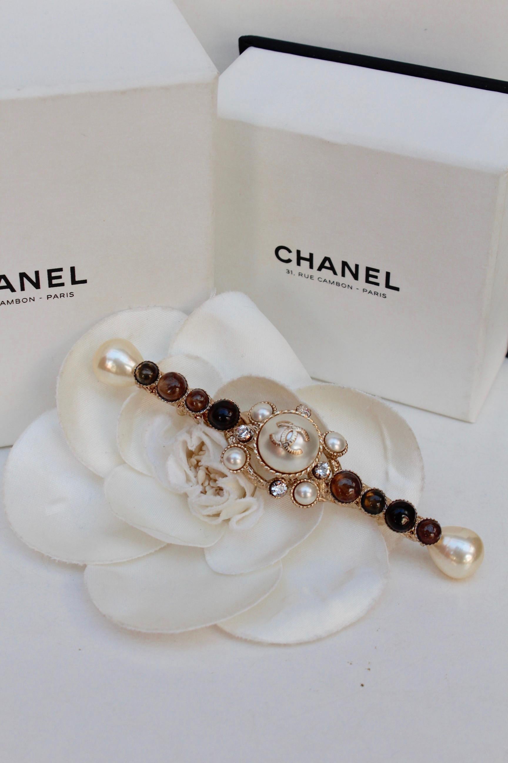 CHANEL (Made in France)
Stunning long light gilted metal brooch with brown, dark brown and pearly glass paste cabochons, decorated with rhinestones. The middle of the piece is made of a pearly rounded cabochon, centered by a light gilted metal