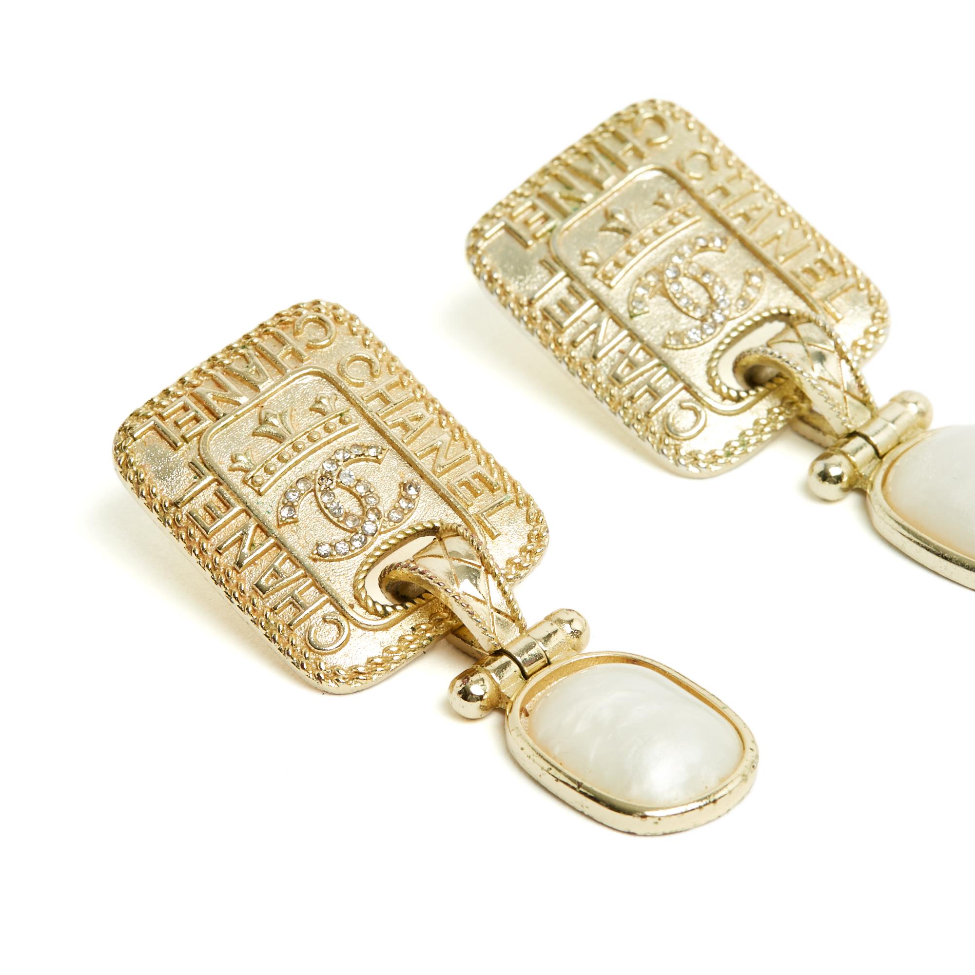 Orecchini Chanel della collezione AH2020 in metallo placcato oro, composti da una piastrina circondata dal nome Chanel e centrata dal simbolo CC incrostato di micro strass bianchi e da un pendente decorato con un cabochon in resina bianca perlata.