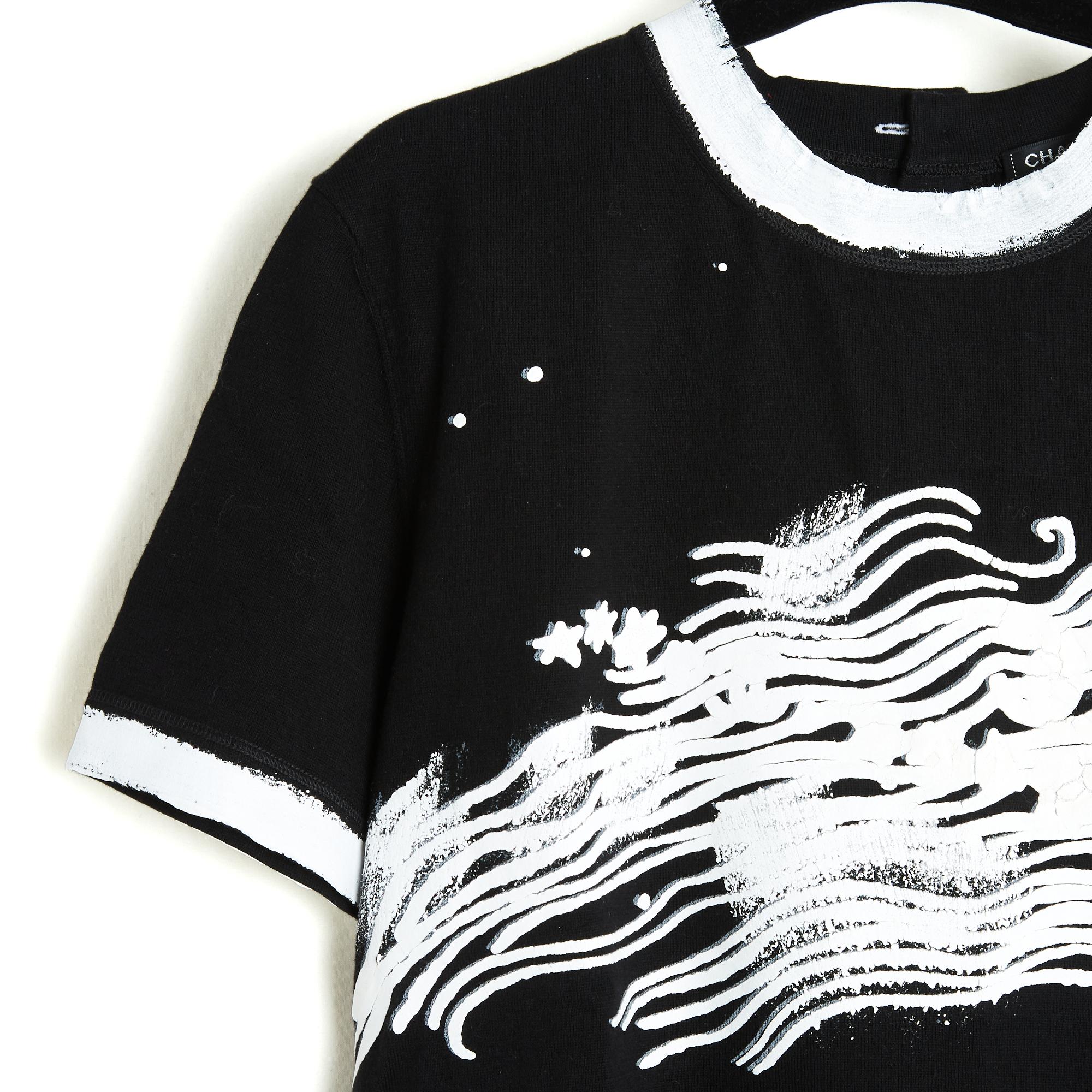 Oberteil aus der Kollektion Chanel Cruise 2022: dickes schwarzes Baumwoll-T-Shirt mit Rundhalsausschnitt (mit drei Logoknöpfen zu schließen), weiß lackierten Manschetten und Saum sowie einem weiß lackierten und gecrackten Motiv auf der Brust. Die