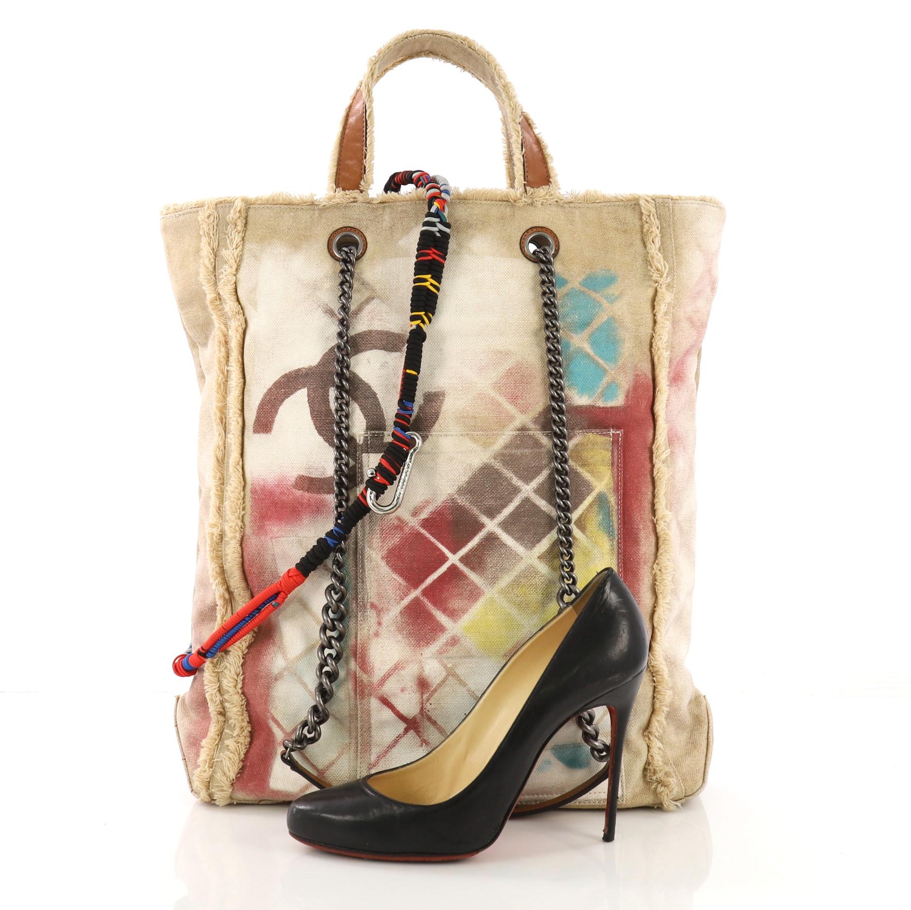 This Chanel Art School Oh My Boy Tote Graffiti Canvas, crafted from beige graffiti printed and sprayed canvas, features dual flat top handles, frayed canvas trim, chain link shoulder straps with leather pads, dangling and multicolored ropes and