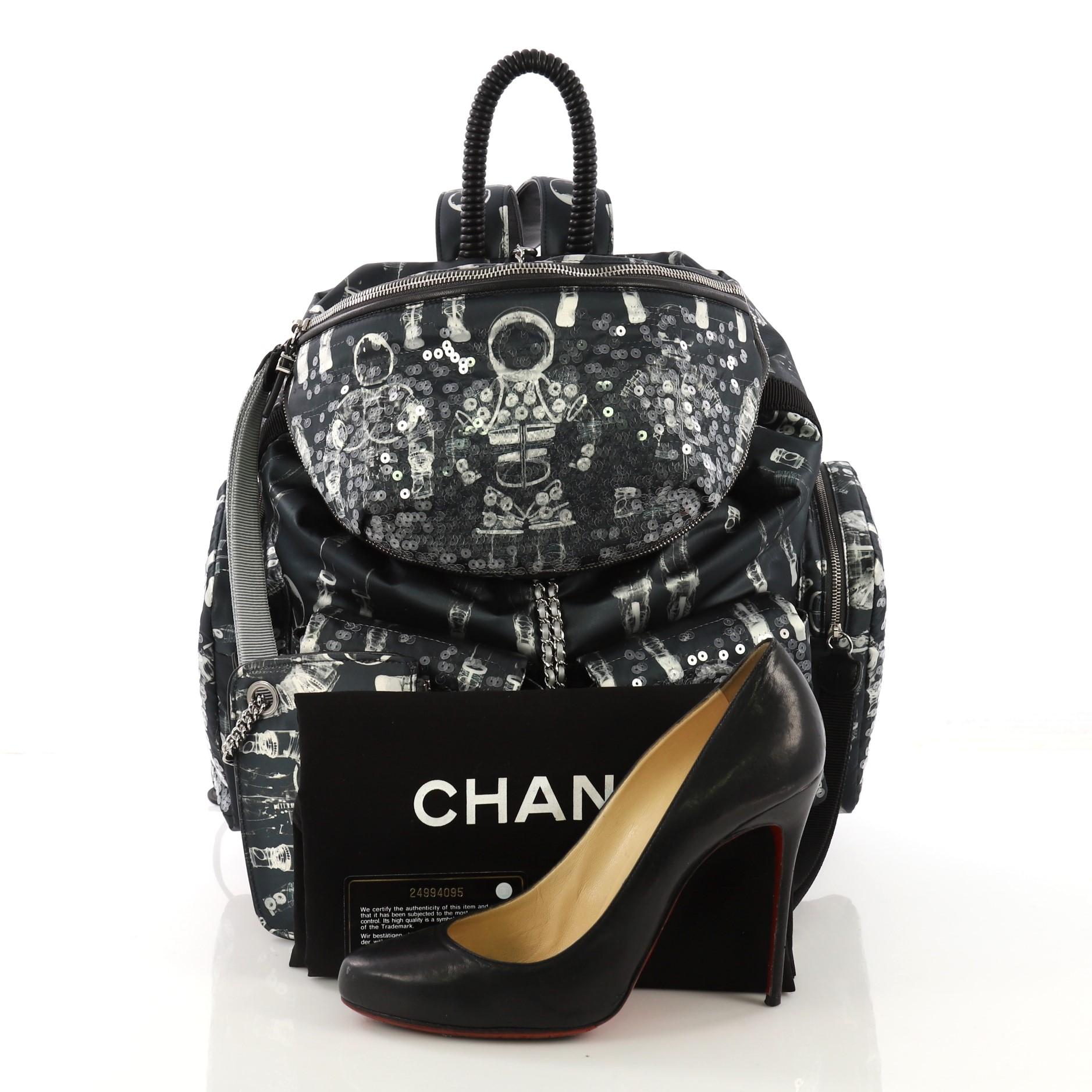 This Chanel Astronaut Essentials Backpack Sequin Embellished Printed Nylon Large, crafted from blue printed nylon, features a rolled top handle, dual flat shoulder straps, leather trim, five exterior pockets, sequin accents, and silver-tone