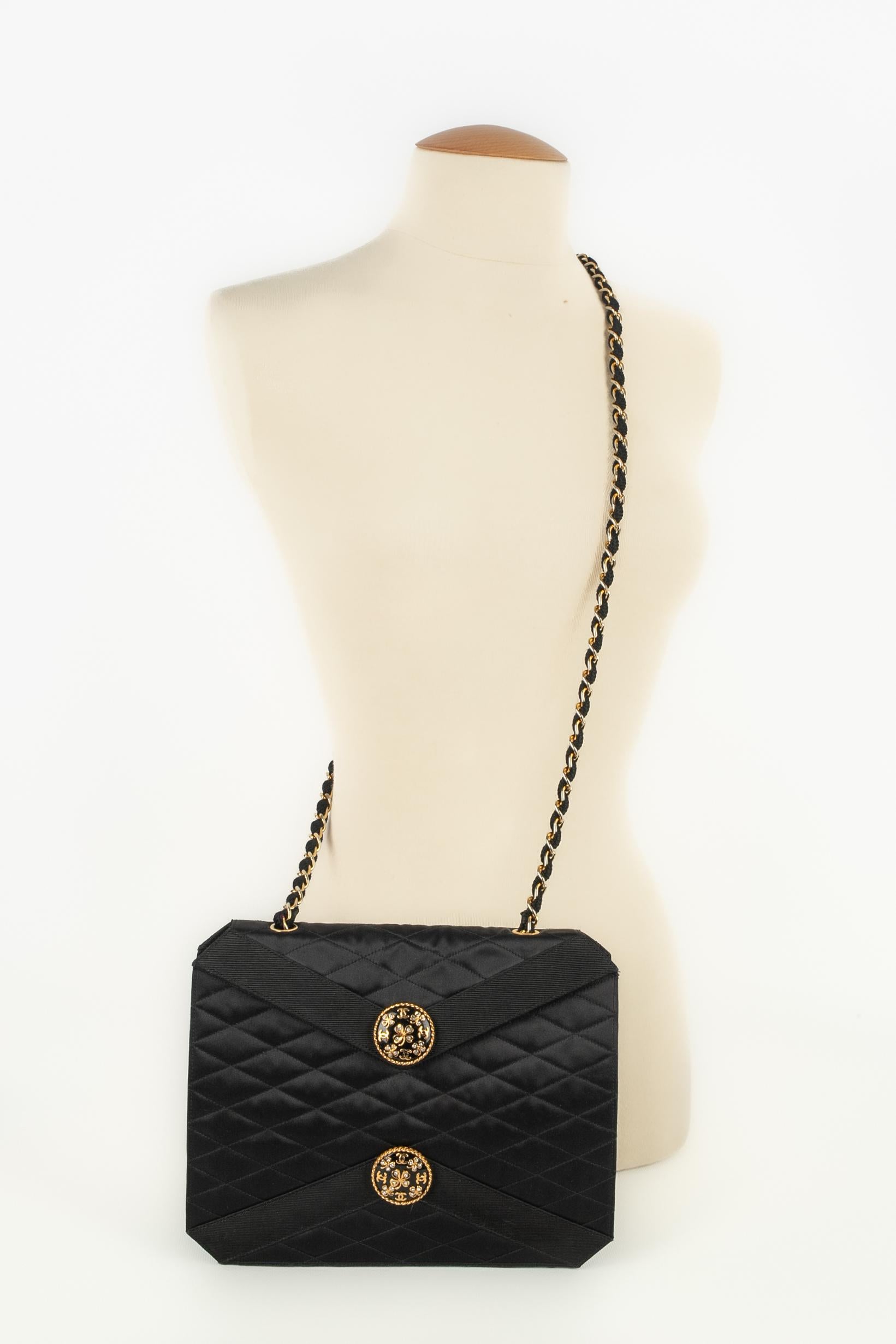 CHANEL - Sac en satin noir matelassé garni de gros grains. Il est orné de deux motifs circulaires décorés de trèfles et d'initiales en strass. Fermeture par bouton pression sur un rabat, anse en chaîne entrelacée. Hologramme 0517841.

Condit :
Très