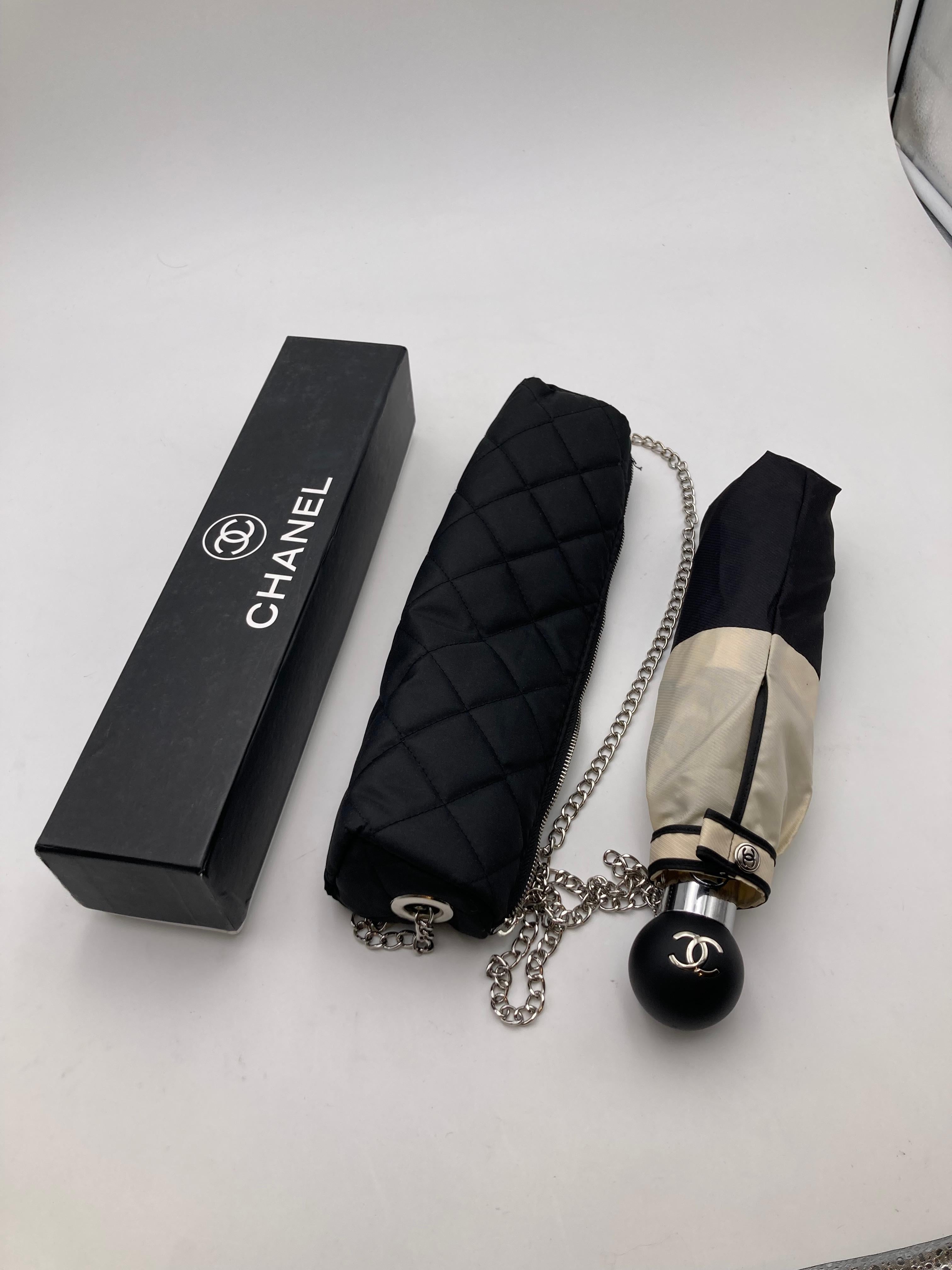 Schöne und exklusive Chanel Regenschirm in schwarz und beige Farbe. Inklusive Schutzhülle und schwarzer gesteppter Stofftasche/Koffer mit silberner Metallkette zum Mitnehmen. Schwarzer Kugelgriff mit CC-Logo. Druckknopf am Griff zum Öffnen.
