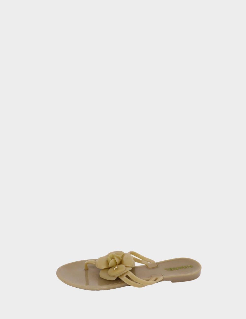 Aggiungi un tocco di giocosa eleganza al tuo guardaroba estivo con questi sandali in gelatina beige di Chanel. Disegnati con una classica infradito, questi sandali presentano l'iconica applicazione del fiore della Camelia, ornata da un sottile logo