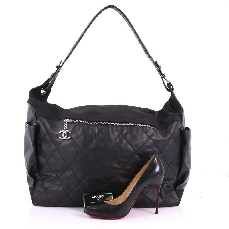 This Chanel Biarritz Hobo Quilted Coated Canvas XL, crafted in black diamond quilted coated canvas, features a looping leather handle, side pockets, front zip pocket with CC charm, and silver-tone hardware. Its top zip closure opens to a black nylon