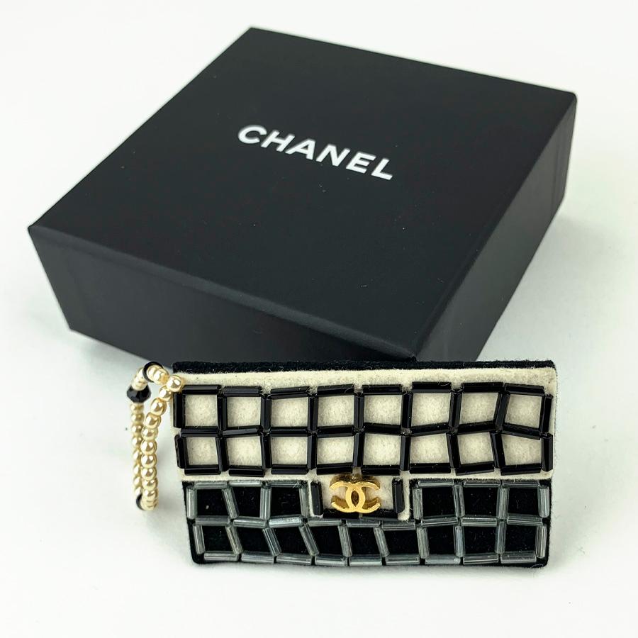 The brooch is signed CHANEL. It represents a CHANEL bag in black and white felt, embroidered with cylindrical beads. The CC clasp is made of gilt metal and the handle of the bag consists of white and black pearls.
The brooch is in very good
