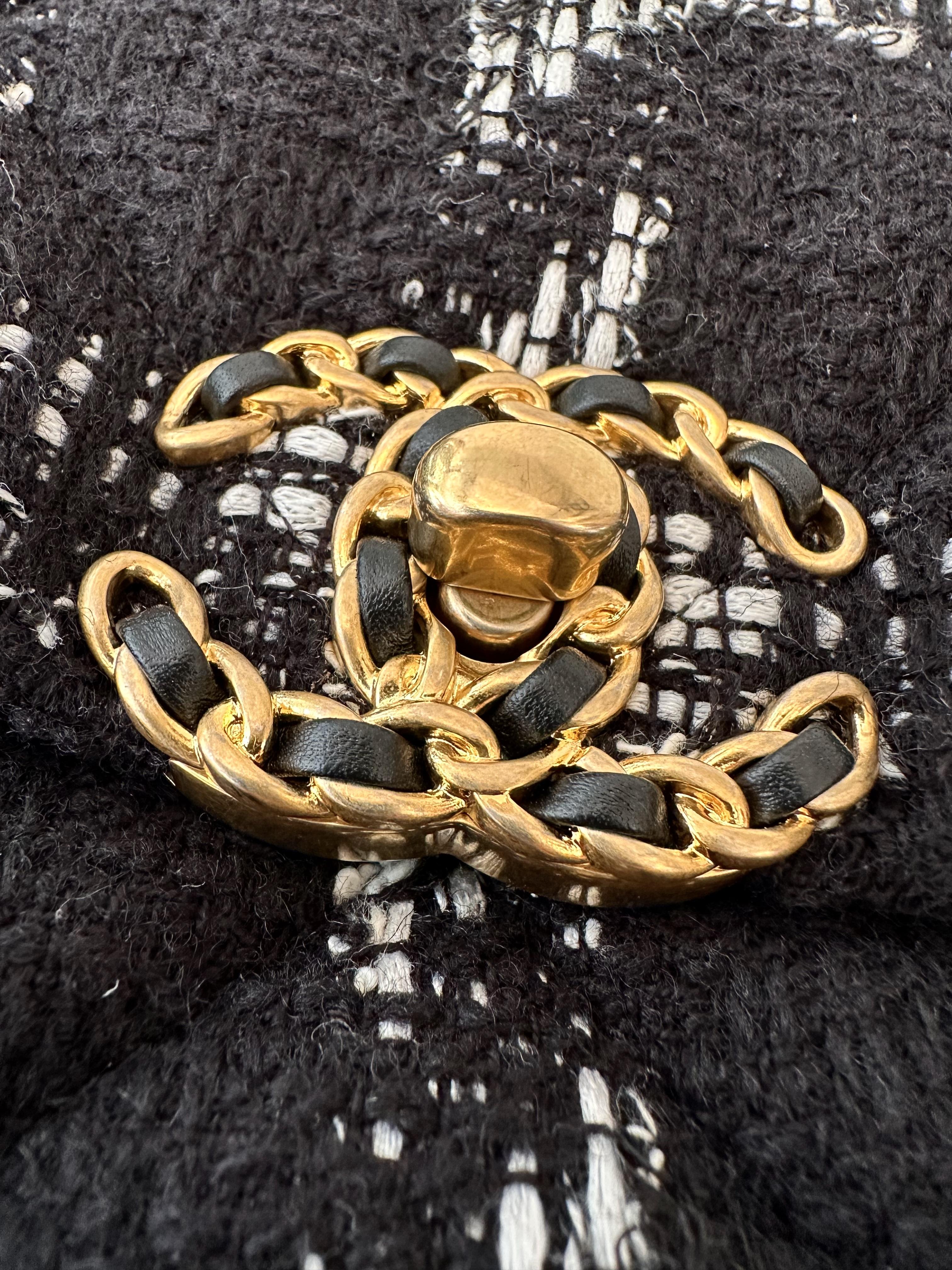 Ce grand sac à rabat Chanel 19 d'occasion est confectionné en tweed matelassé noir avec un motif blanc en forme de losange. 
Il est doté d'une poignée supérieure en chaîne dorée de 10 cm (environ 3,9