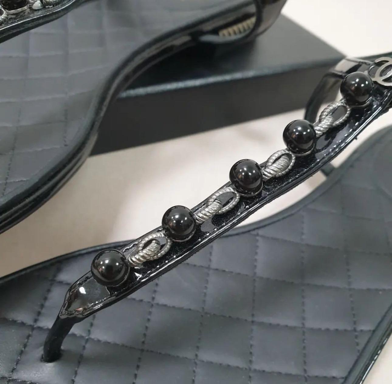 CHANEL Patent Perlen Kette Zehensteg Sandalen in Schwarz. 
Diese modischen Zehenstegsandalen sind aus schwarzem Lackleder mit gesteppten Lammfellsohlen gefertigt. 
Der Riemen ist mit der für Chanel typischen Perlenkette verziert. 
Sie verfügen über