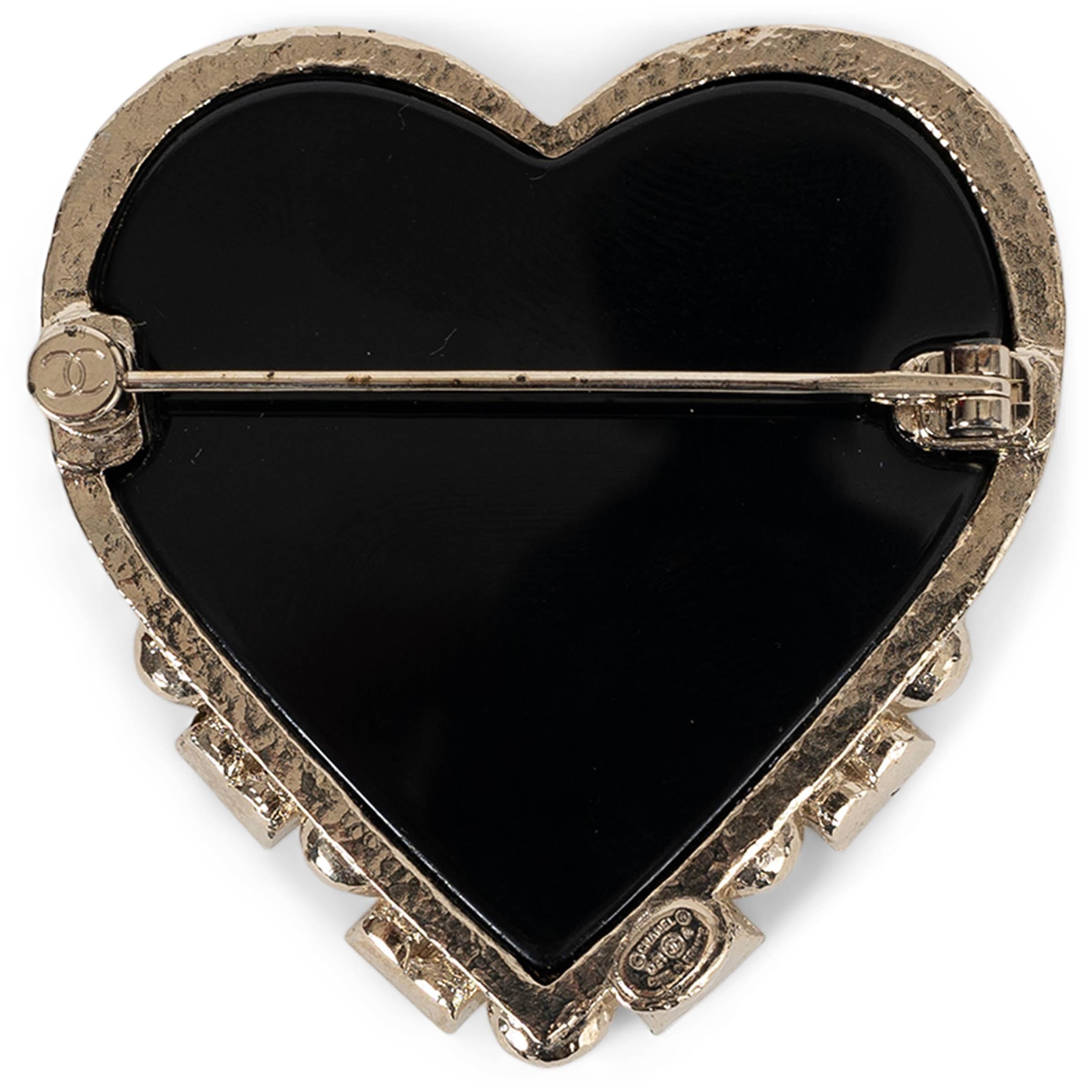 This 100% authentic Chanel heart brooch in black resin is a striking statement piece, blending bold contrast with refined elegance. Encased in a beige gold-tone metal frame adorned with shimmering crystals, it radiates a luxurious touch. A signature