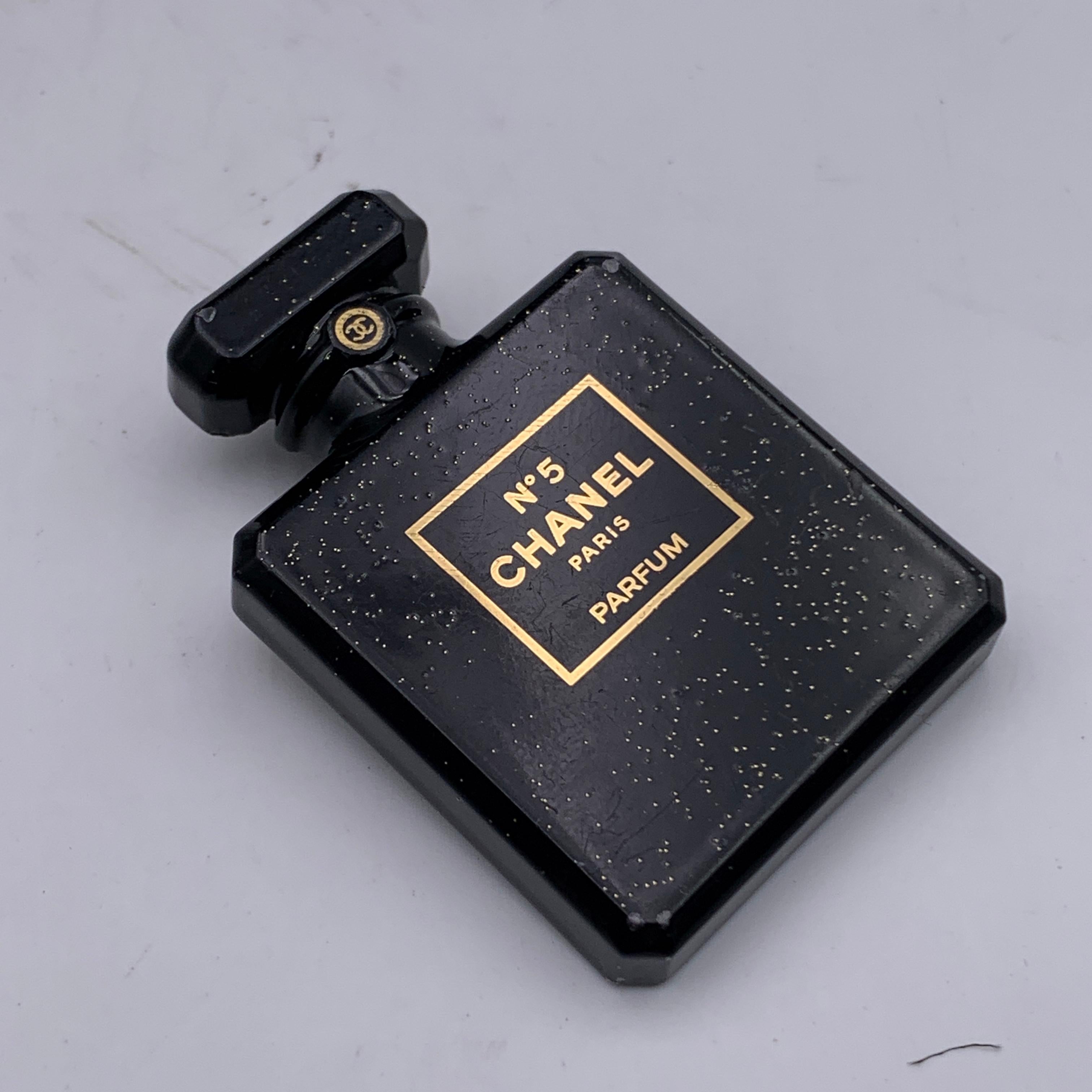 Chanel black brooch from the 2021 Fall/Winter collection, shaped as the iconic Chanel N°5 Parfum bottle. Black resin accented with gold metal glitter. Safety pin closure. 'Chanel - U21 CC K - Made in France' oval hallmark on the back. Measurements: