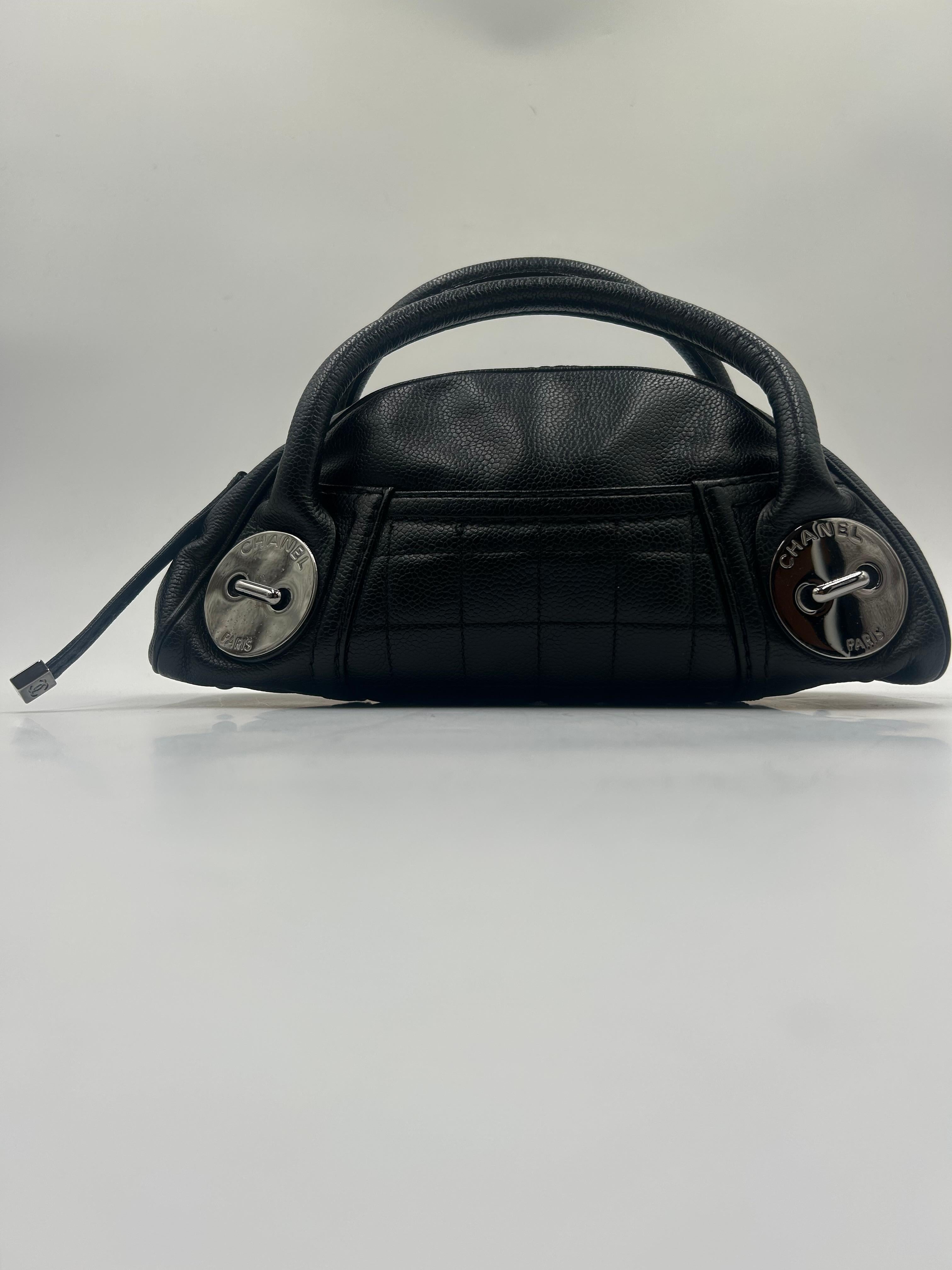 This Chanel Black Leather Button Satchel, showcases a sophisticated and slightly vintage-inspired design. It is crafted from smooth black leather with a structured silhouette and subtle shine. 
The body has minimal quilting, emphasizing clean lines