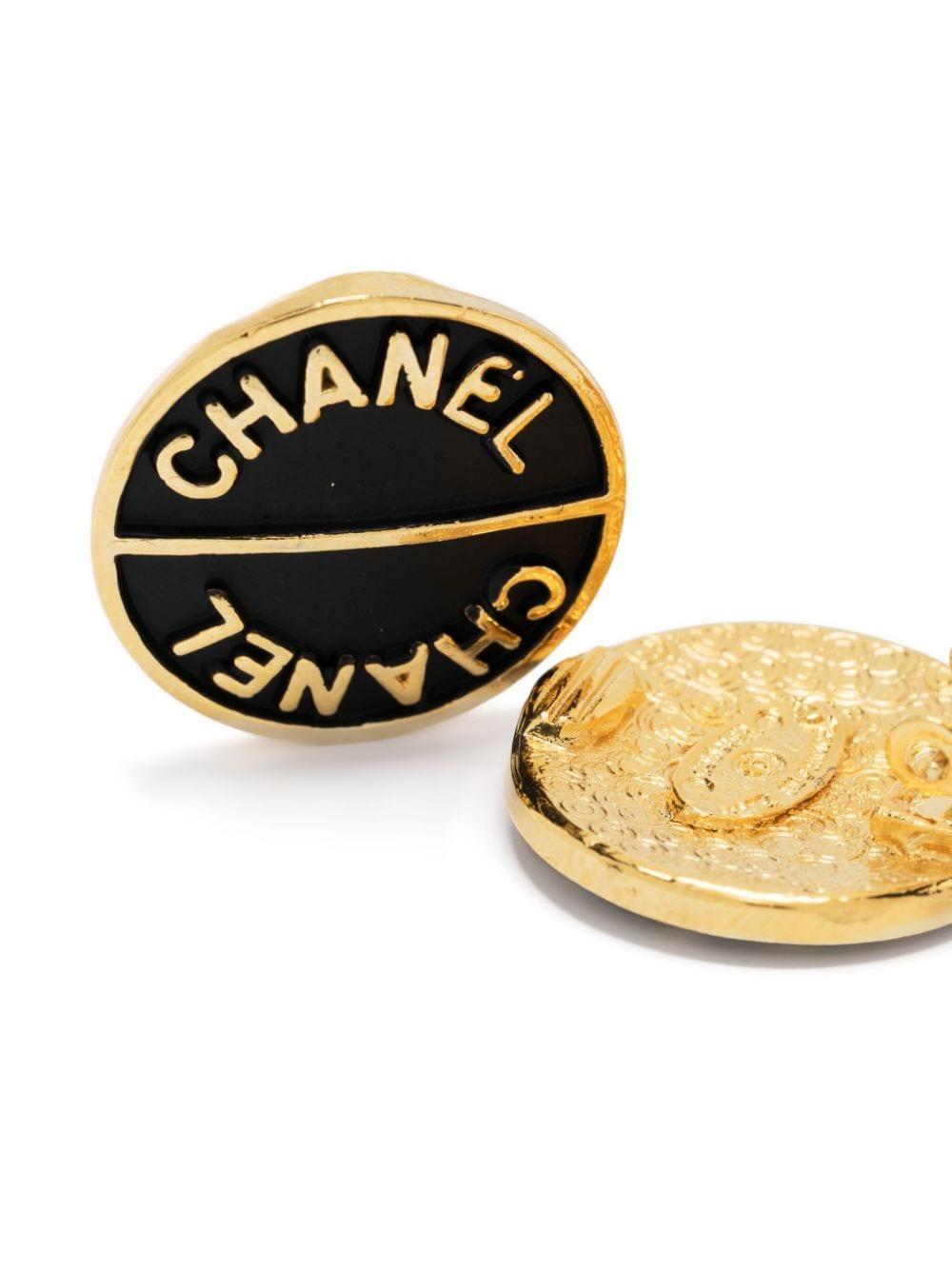 Schwarze Chanel-Ohrringe mit Logo-Prägung aus den 1990er Jahren, mit folgenden Eigenschaften: signiertes Logo, goldfarbene Umrandung, geprägtes Logo auf der Vorderseite, Clip-Verschluss, entsteinte Plakette auf der Rückseite. 
Circa 1990er