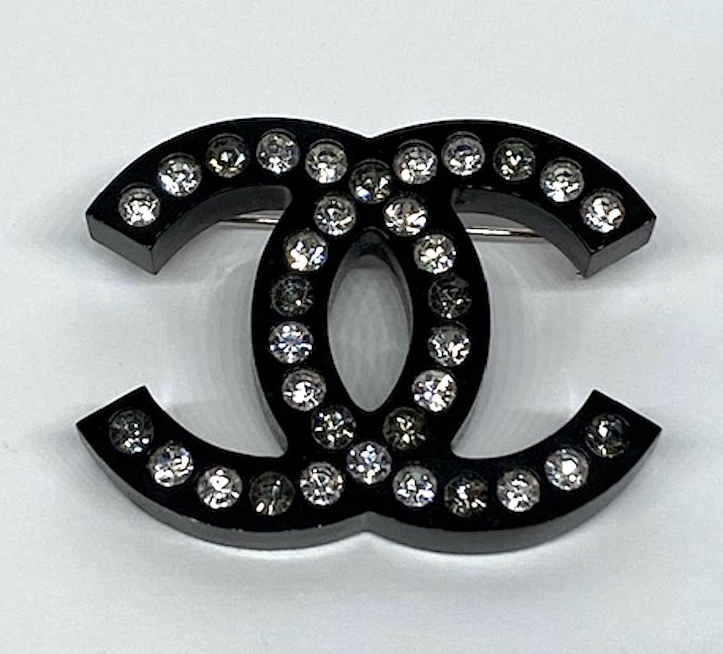 From the Spring Collection 2016 is this black lucite iconic Chanel interlocking CC logo pin set with clear and grey rhinestones. The black lucite is pin is 2 inches wide, 1.5 inches high and .25 on an inch thick. With the rhodium pin backing it is