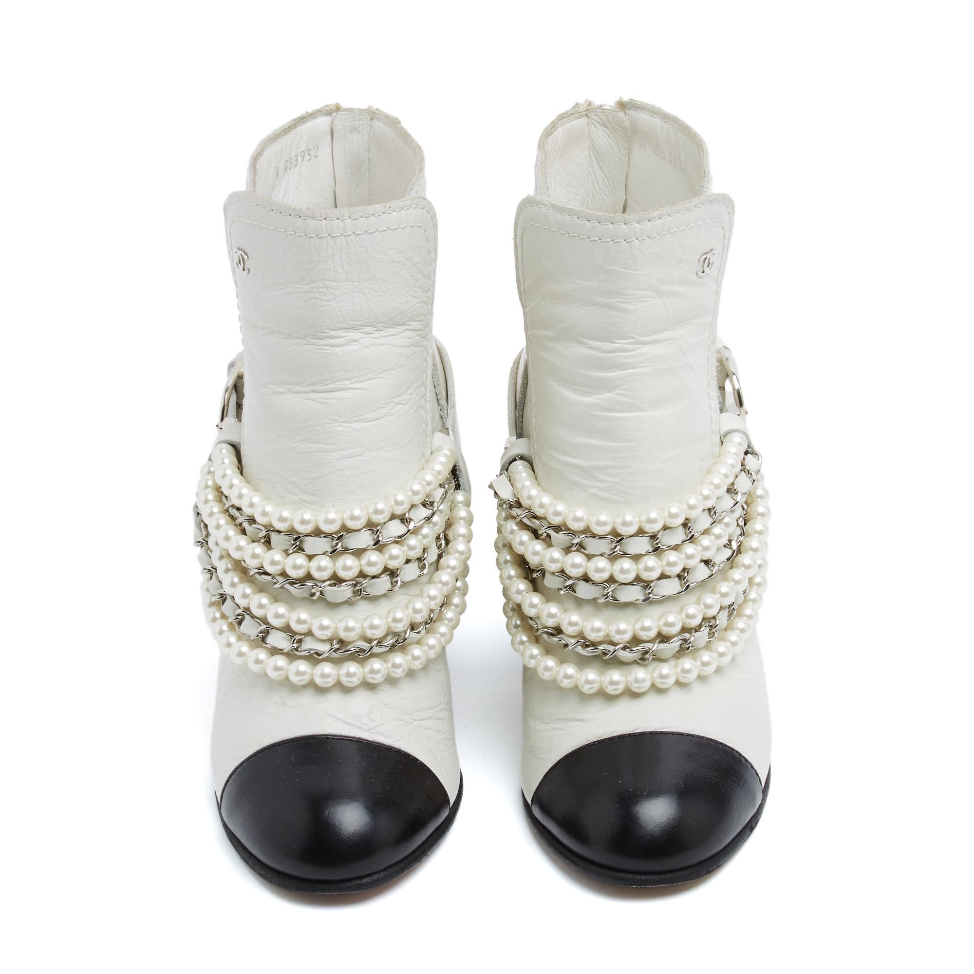 Chanel ankle boots in white leather, with a leather-covered heel and a round black leather toe. The straps are adorned with rows of decorative pearls alternating with rows of silver-tone metal chain interwoven with white leather. They fasten with a