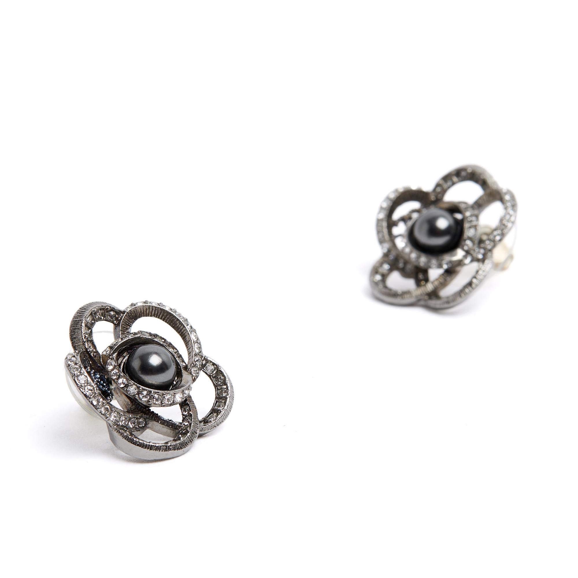 Orecchini Chanel con un motivo a camelia stilizzato incrostato di strass bianchi e grigi, un piccolo logo CC (visibile attraverso uno dei petali) e una perla fantasia al centro. Diametro circa 2,2 cm. Gli orecchini vengono consegnati senza fattura o