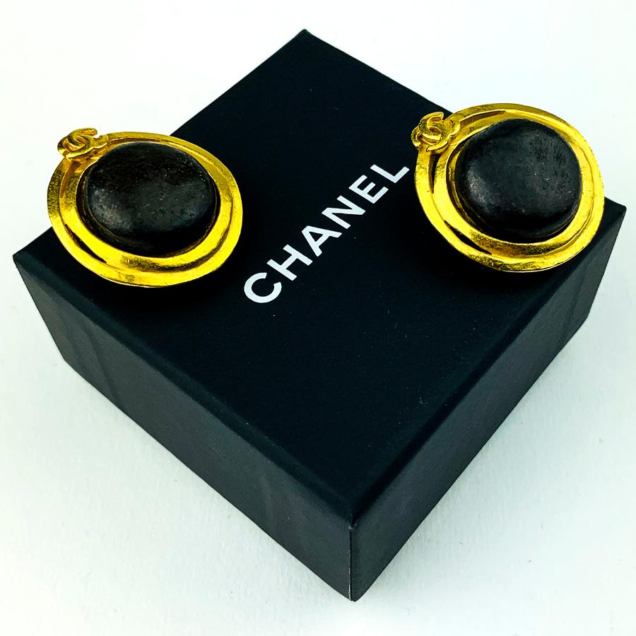 Die Ohrclips stammen von Maison CHANEL. Sie sind rund und haben in der Mitte eine Kugel aus braunem Leder, die von fein vergoldetem Metall mit dem CC-Logo der Marke umgeben ist.
Die Ohrringe sind in sehr gutem Zustand für einen Jahrgang. Die Clips