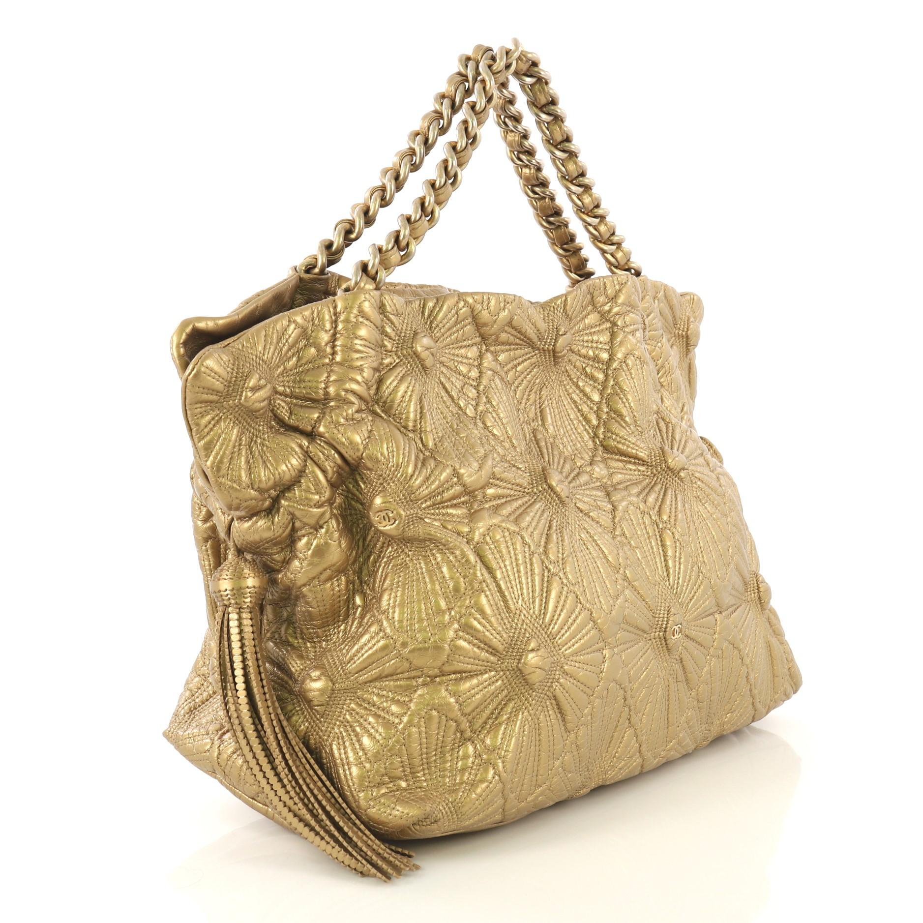 This Chanel Ca D'Oro Tote Quilted Lambskin Large, crafted from gold metallic quilted lambskin, features dual woven-in leather chain handles, side tassels, floral embroidery design, and gold-tone hardware. Its magnetic snap closure opens to a tan