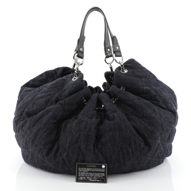 This Chanel Cabas Tote Quilted Denim Large, crafted in blue denim, features leather straps with silver chain links threaded through eyelets that cinches the stretch fabric top of the bag, a large stitched frontal Chanel CC logo, and silver-tone