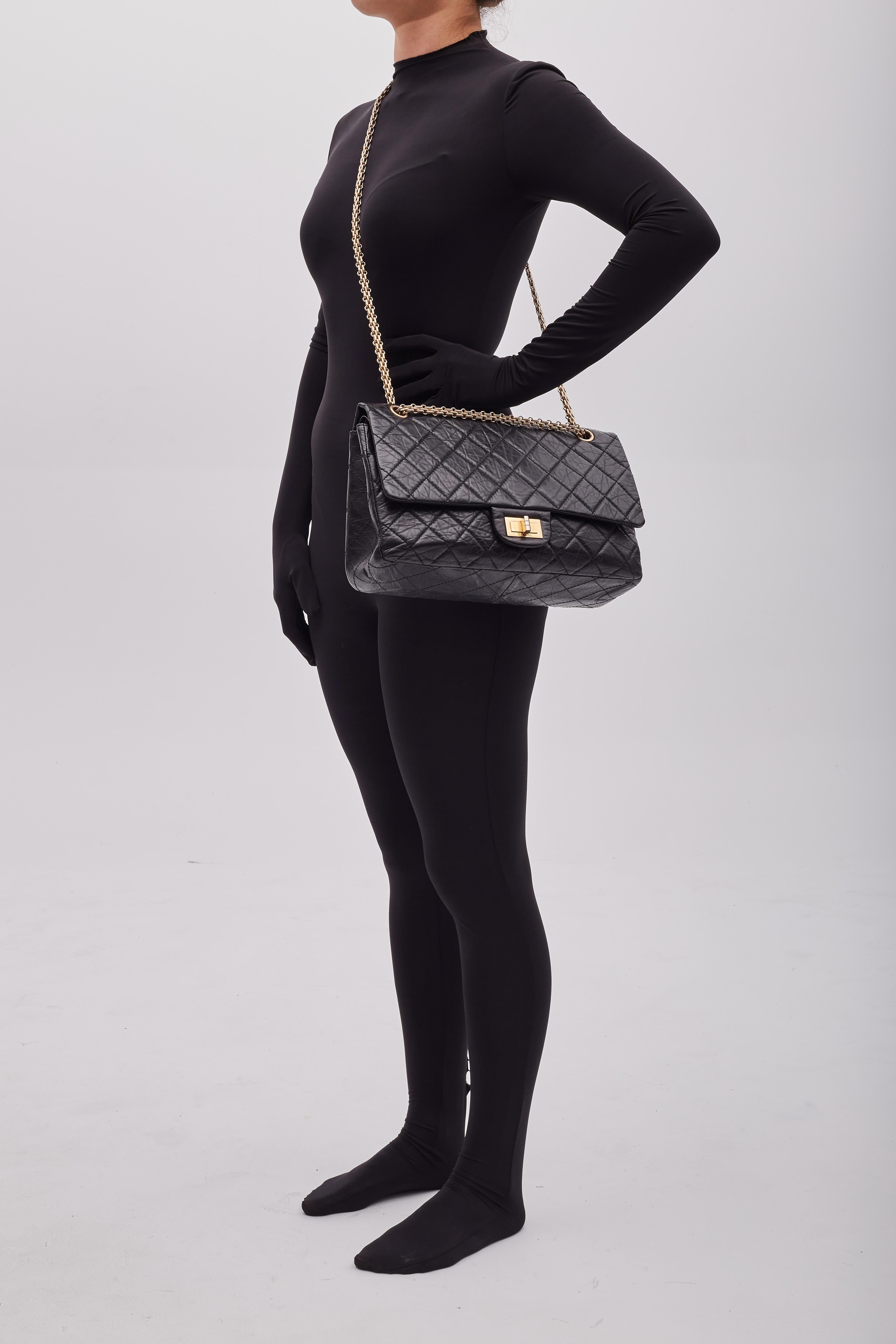 Voici le sac à rabat Chanel Reissue 2.55 227 dans la plus grande taille de ce modèle. Le sac est confectionné en cuir de veau vieilli matelassé de couleur noire avec des ferrures de tonalité dorée antiquaire. Le sac est doté d'une bandoulière