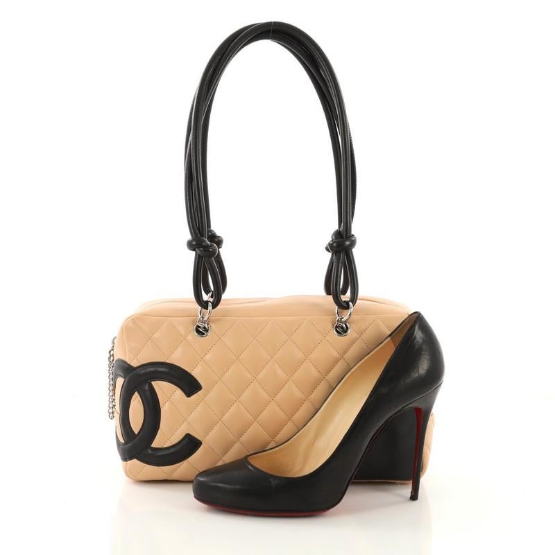 This Chanel Cambon Bowler Bag Quilted Leather Medium, crafted from beige diamond quilted leather, features a large black interlocking CC side logo, rolled leather handles with knotted ends, and silver-tone hardware. Its zip closure opens to a black